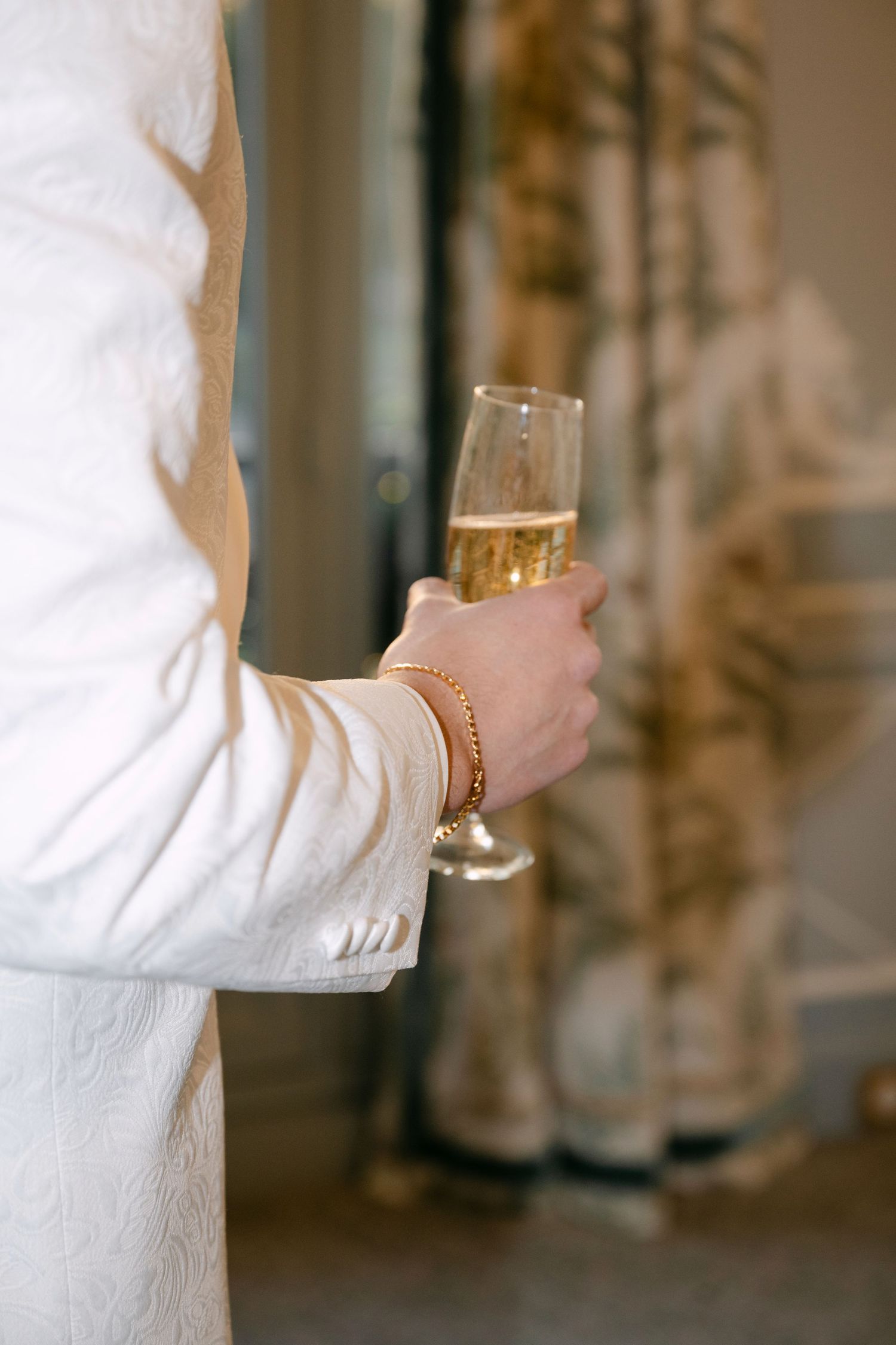 Close-up of person in white suit holding champagne flute with gold bead bracelet at wedding.
