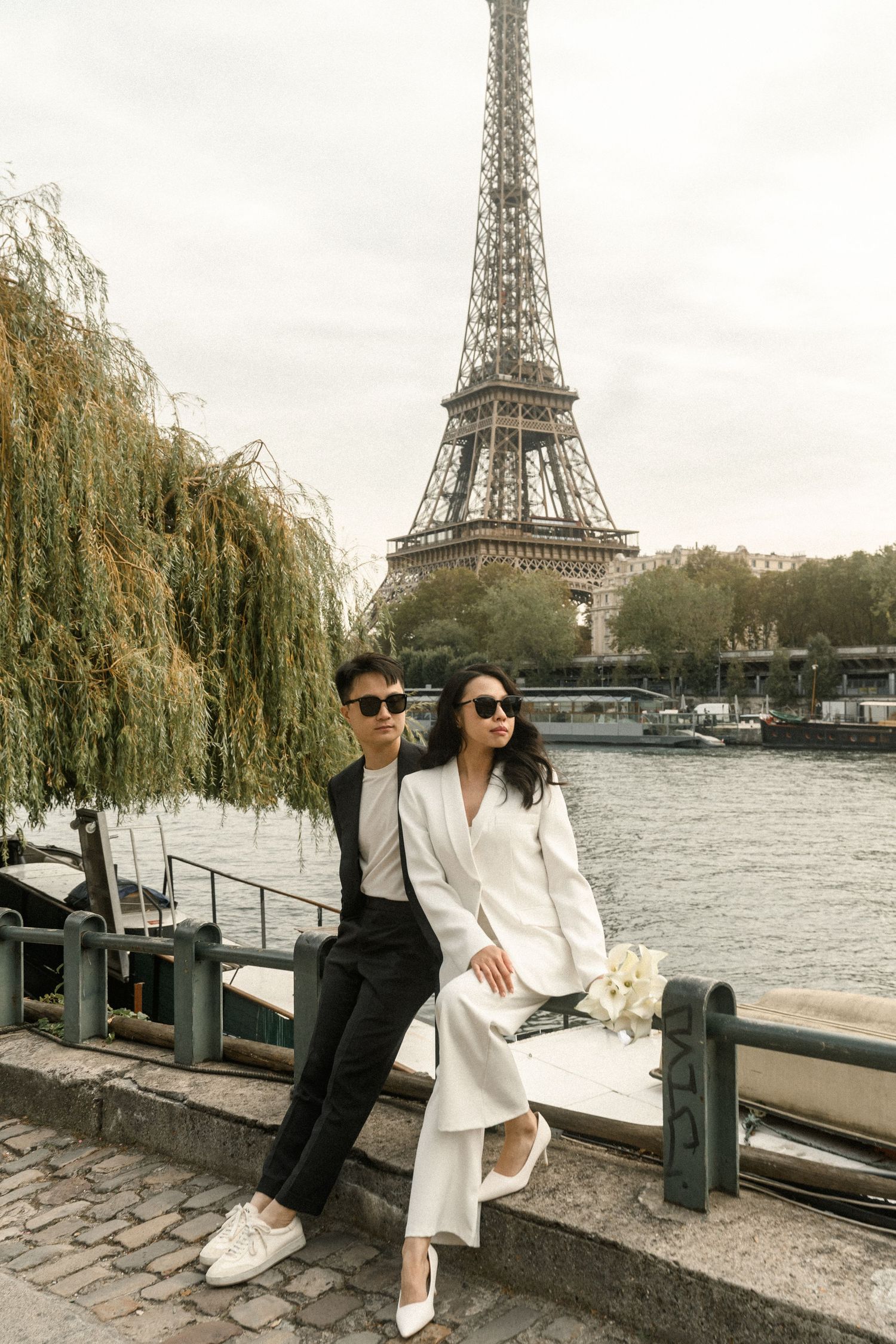 An Asian couple in black and white suits pose together by the Seine River with the Eiffel Tower behind.
