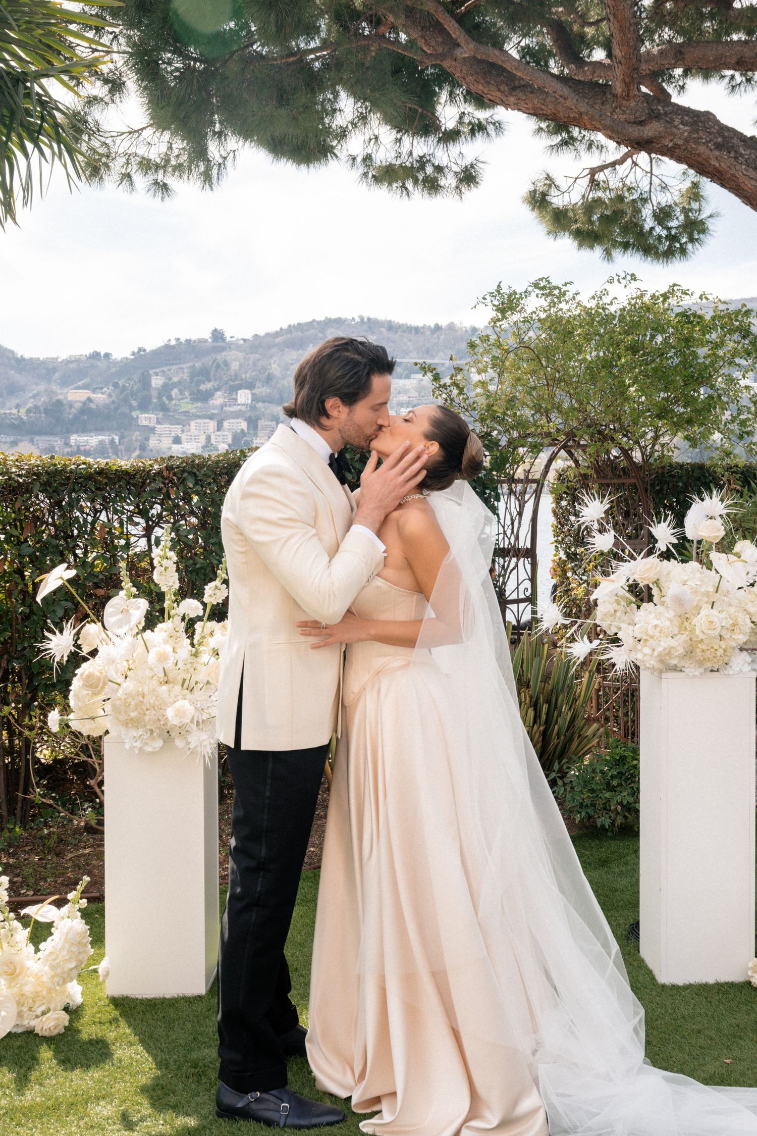 Bride and groom sharing first kiss at outdoor wedding ceremony with white floral arrangements.