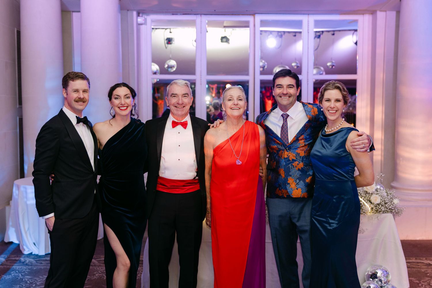 Group of six elegantly dressed wedding guests posing together at a formal reception venue.