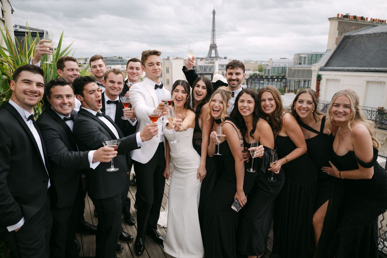 Wedding party group photo with bride and groom holding cocktails on a Paris rooftop with Eiffel Tower view.