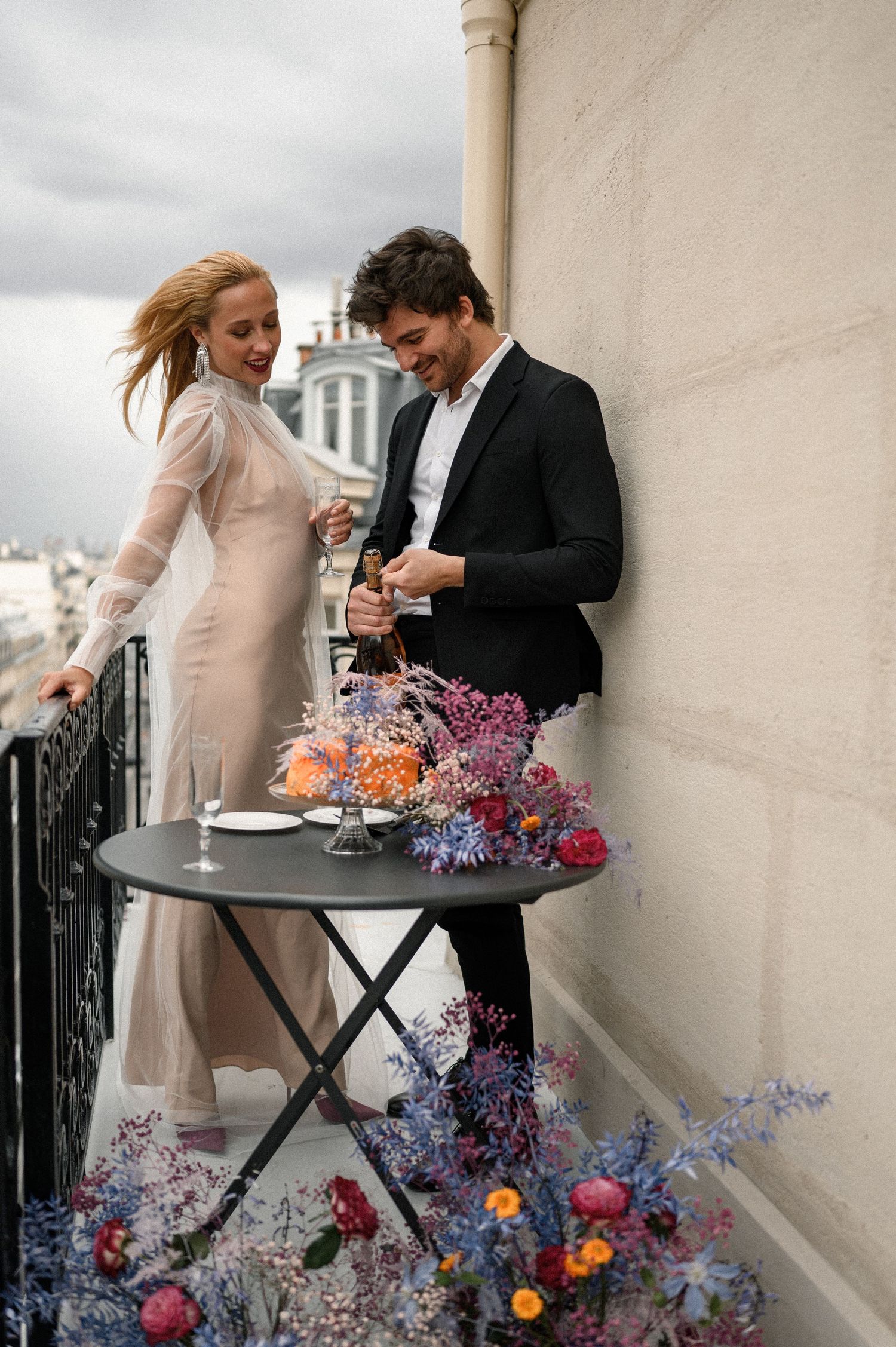 Couple celebrating on a Paris balcony with colorful floral arrangement, bride in sheer gown and groom in black suit.
