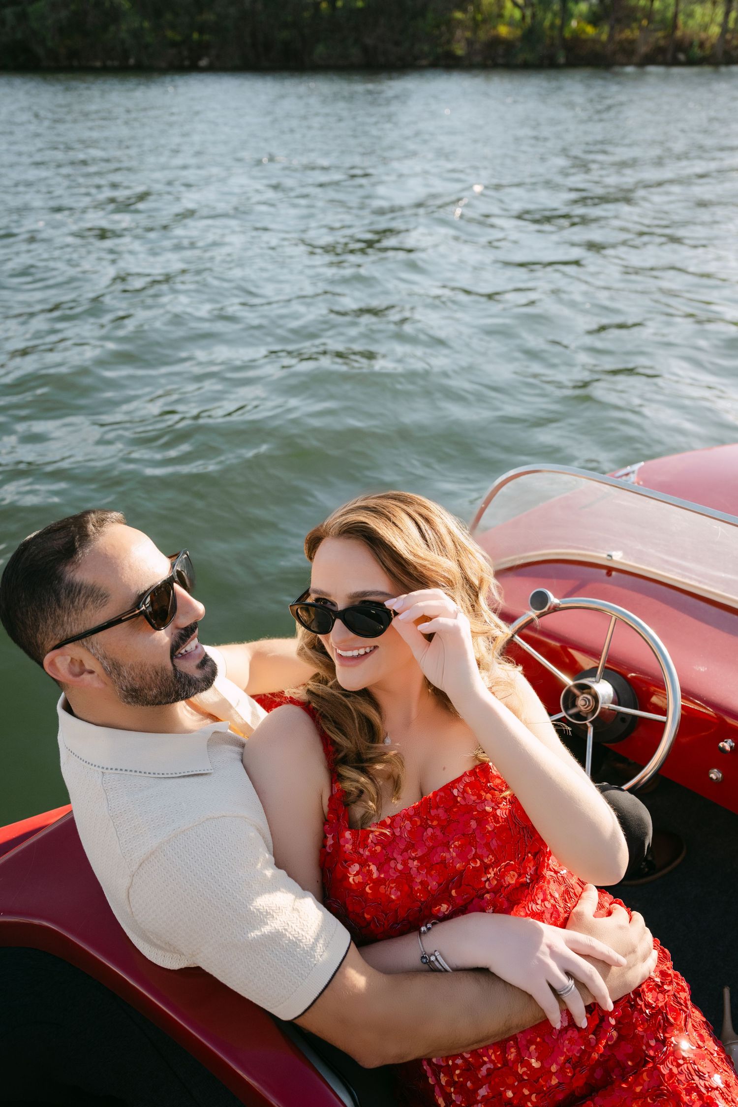Happy couple in sunglasses embracing on a classic red boat on the water, woman in red sequin dress. Austin Wedding Photographer
