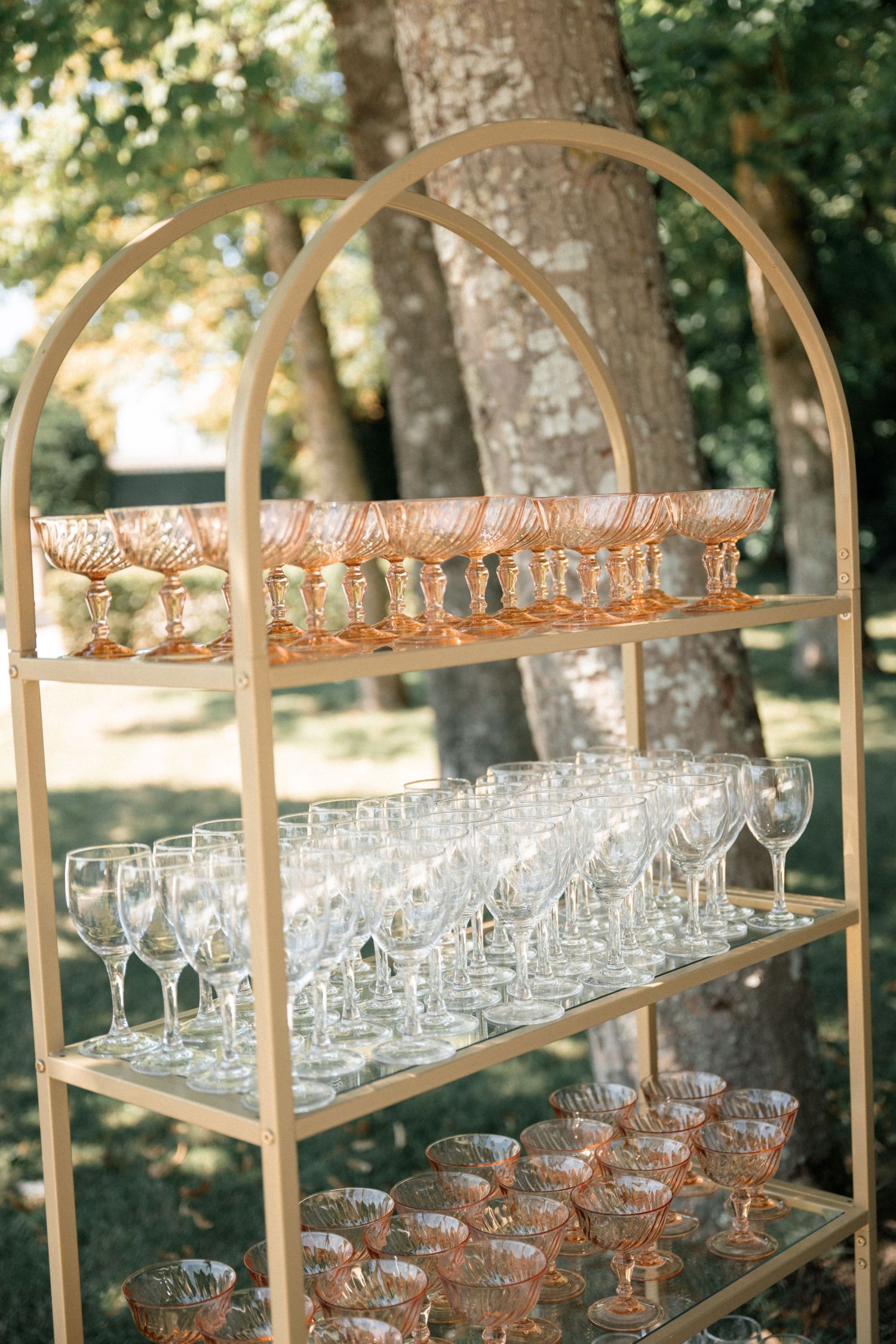 Gold tiered display shelf with pink champagne coupes and clear wine glasses outdoors at an elegant wedding reception.