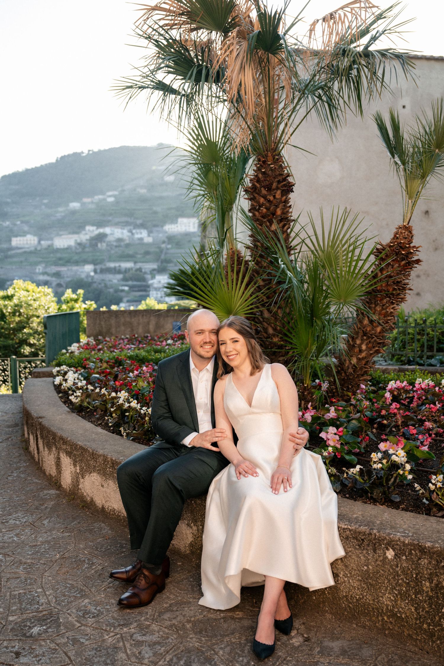 Bride and groom sit together on garden bench with palm trees and mountain views in Italy.