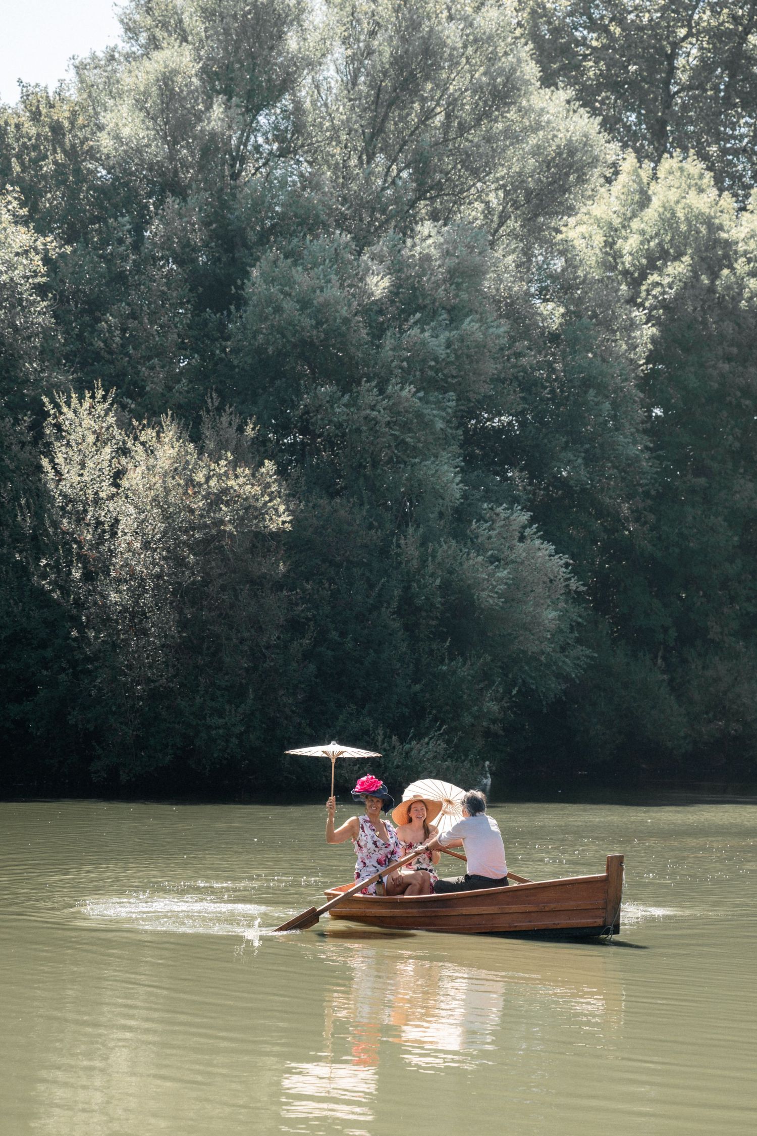 Two people rowing a wooden boat with a parasol on a calm river surrounded by trees.