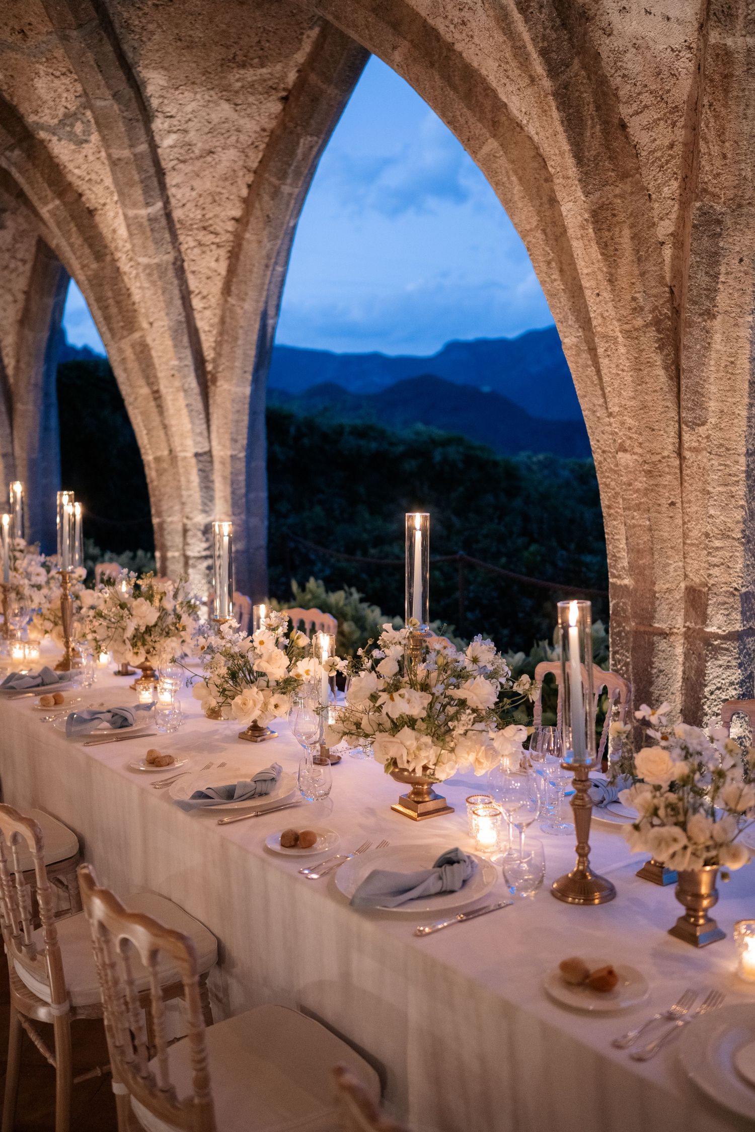Stunning Italian wedding reception table set under gothic stone arches with mountain views at twilight.