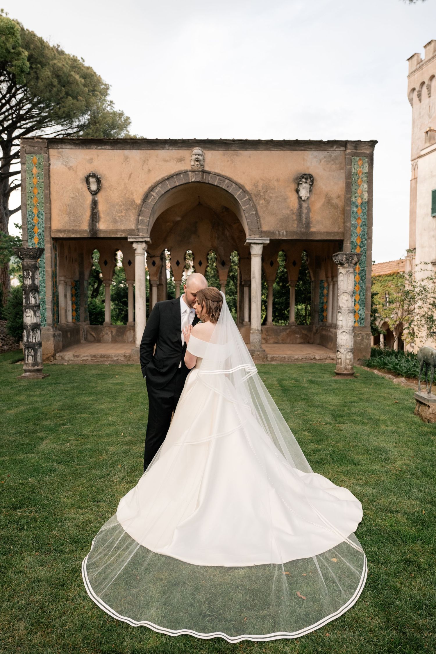 Bride and groom share a kiss in a lush garden beside a historic Italian villa archway on their wedding day.