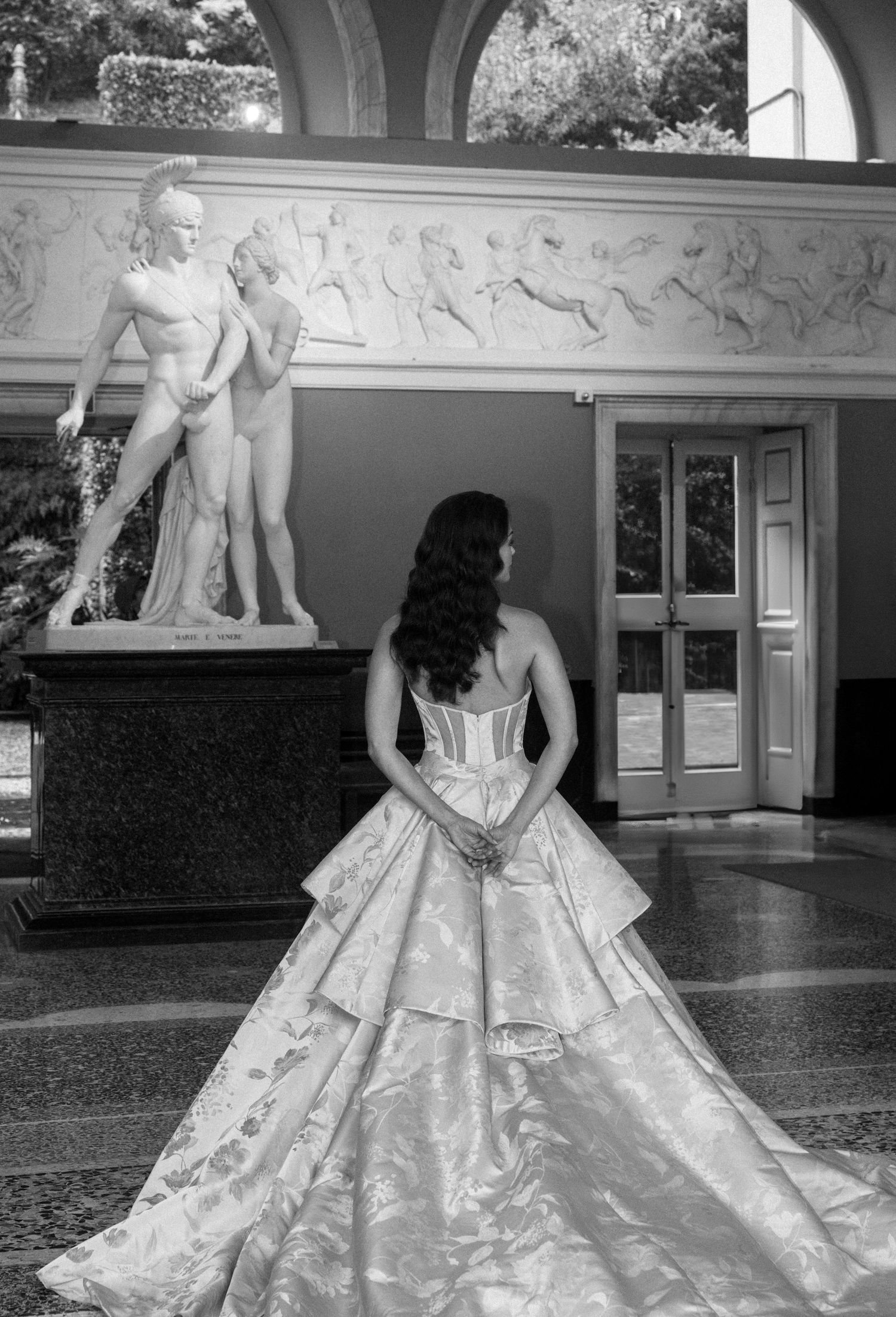 Black and white photo of a bride in an elegant ballgown viewed from behind, standing before classical sculptures in a grand hall.