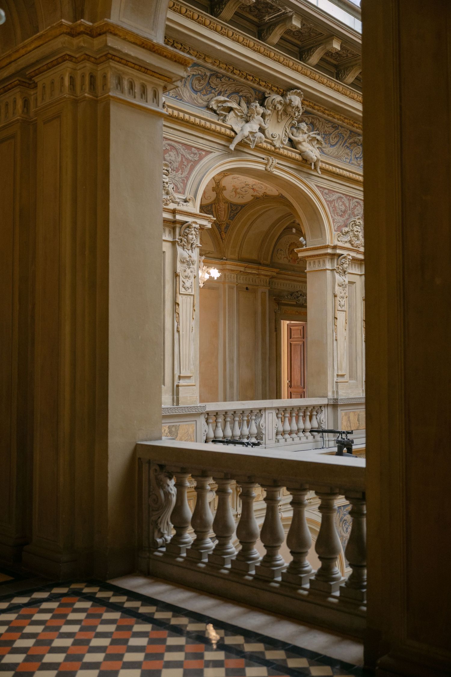 Ornate baroque interior with balustrade, arched doorways, decorative moldings, and checkered marble floor in a grand historic building.