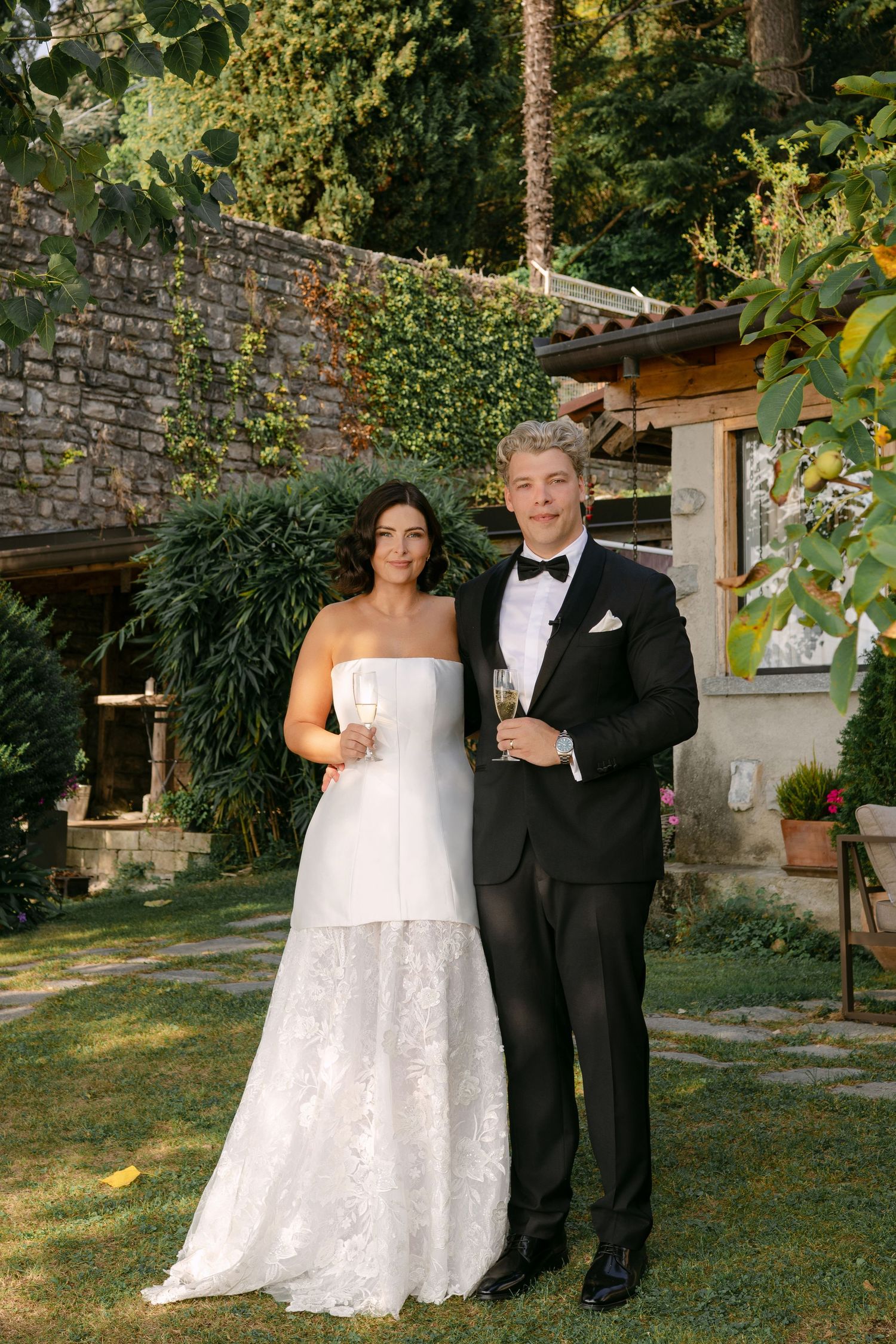 Bride and groom holding champagne glasses pose together outdoors in garden setting, wearing wedding dress and black tuxedo.