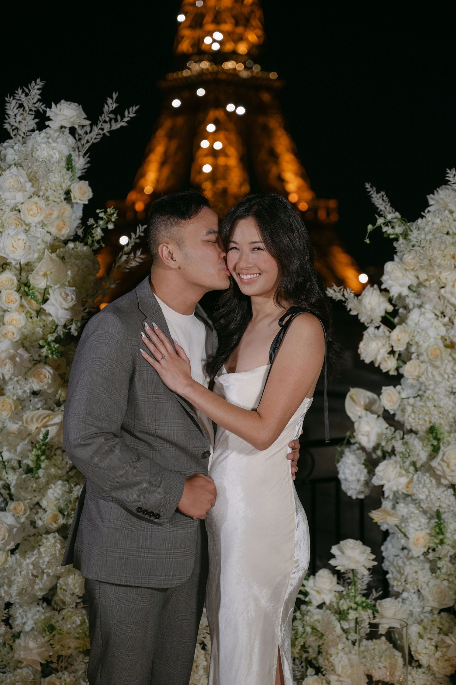 Smiling woman in a white slip dress is kissed by her partner beside white floral arrangements with the illuminated Eiffel Tower behind.