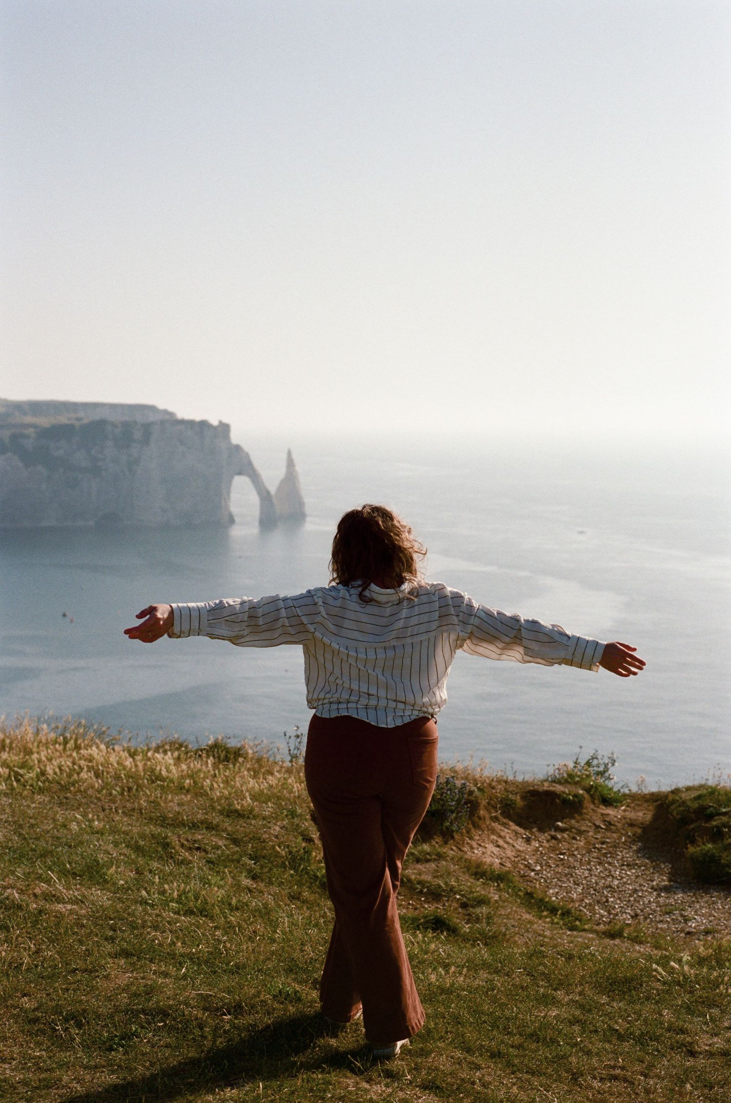 Woman with arms outstretched standing on coastal cliffs overlooking the sea and natural rock arch formation.