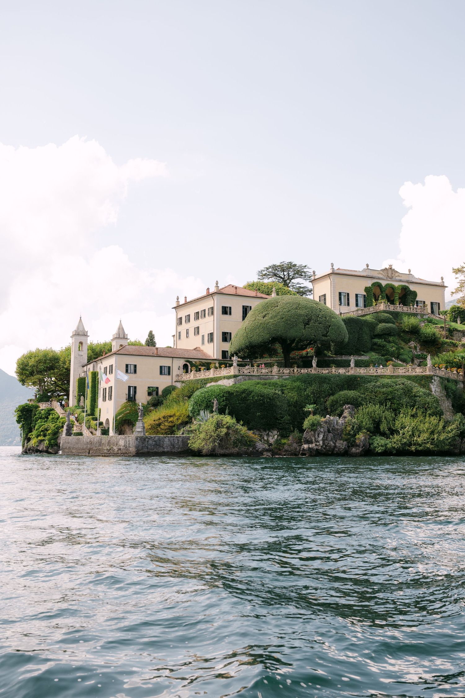 Elegant Italian villa on Lake Como shoreline surrounded by lush green trees and gardens, viewed from the water.