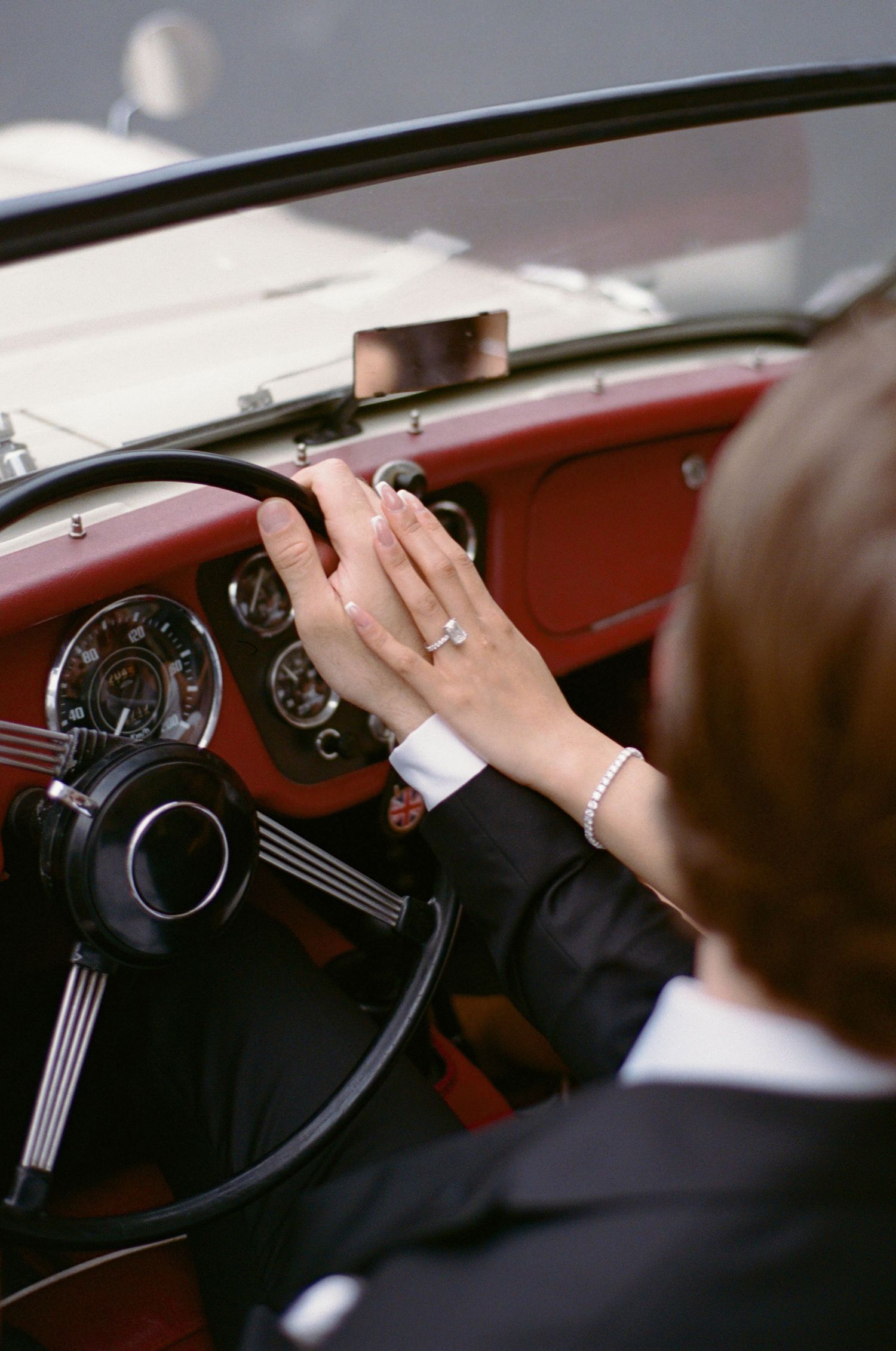 Person wearing a diamond ring and bracelet gripping the steering wheel of a vintage red and white convertible car.