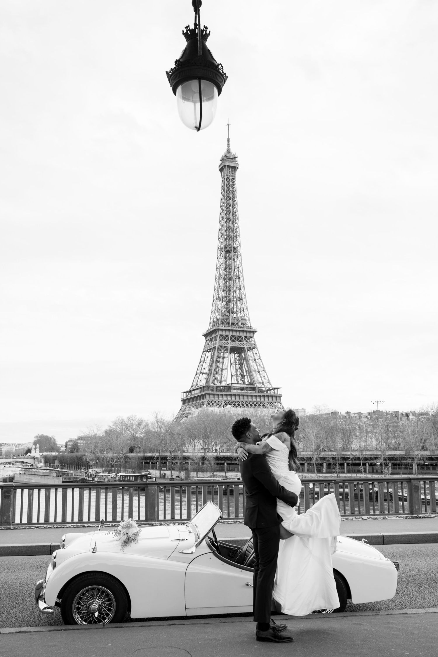 Black and white photo of newlyweds embracing beside vintage white convertible with Eiffel Tower backdrop.