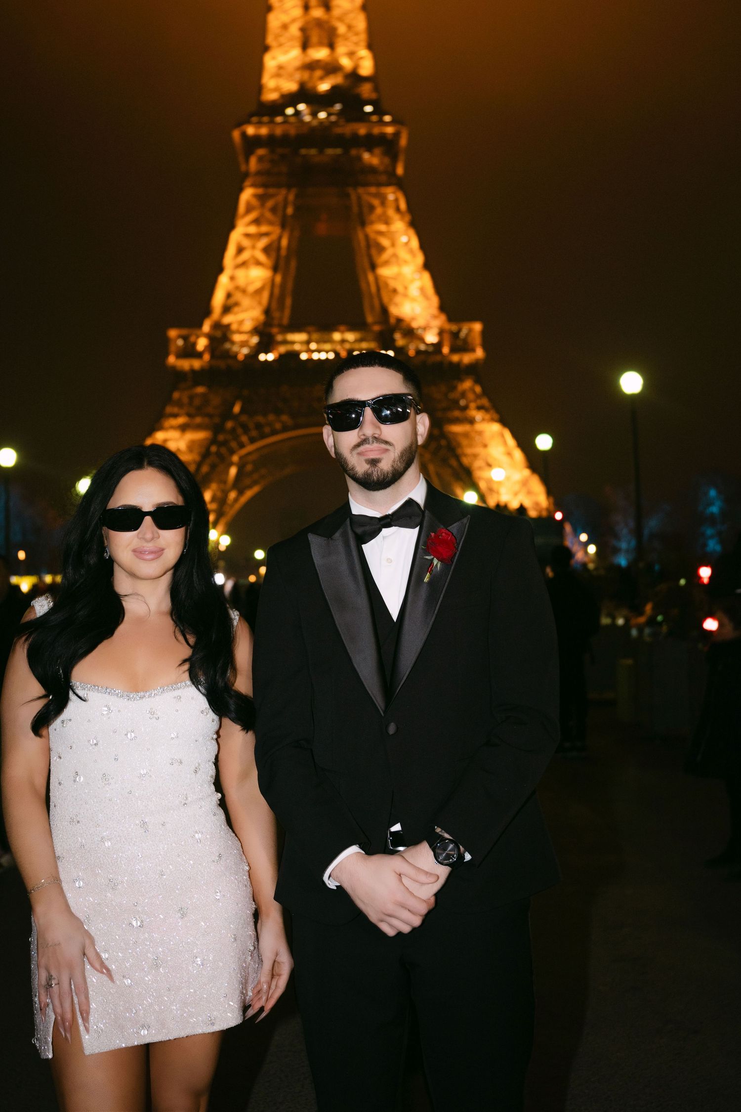 Couple in formal attire and sunglasses posing in front of illuminated Eiffel Tower at night.