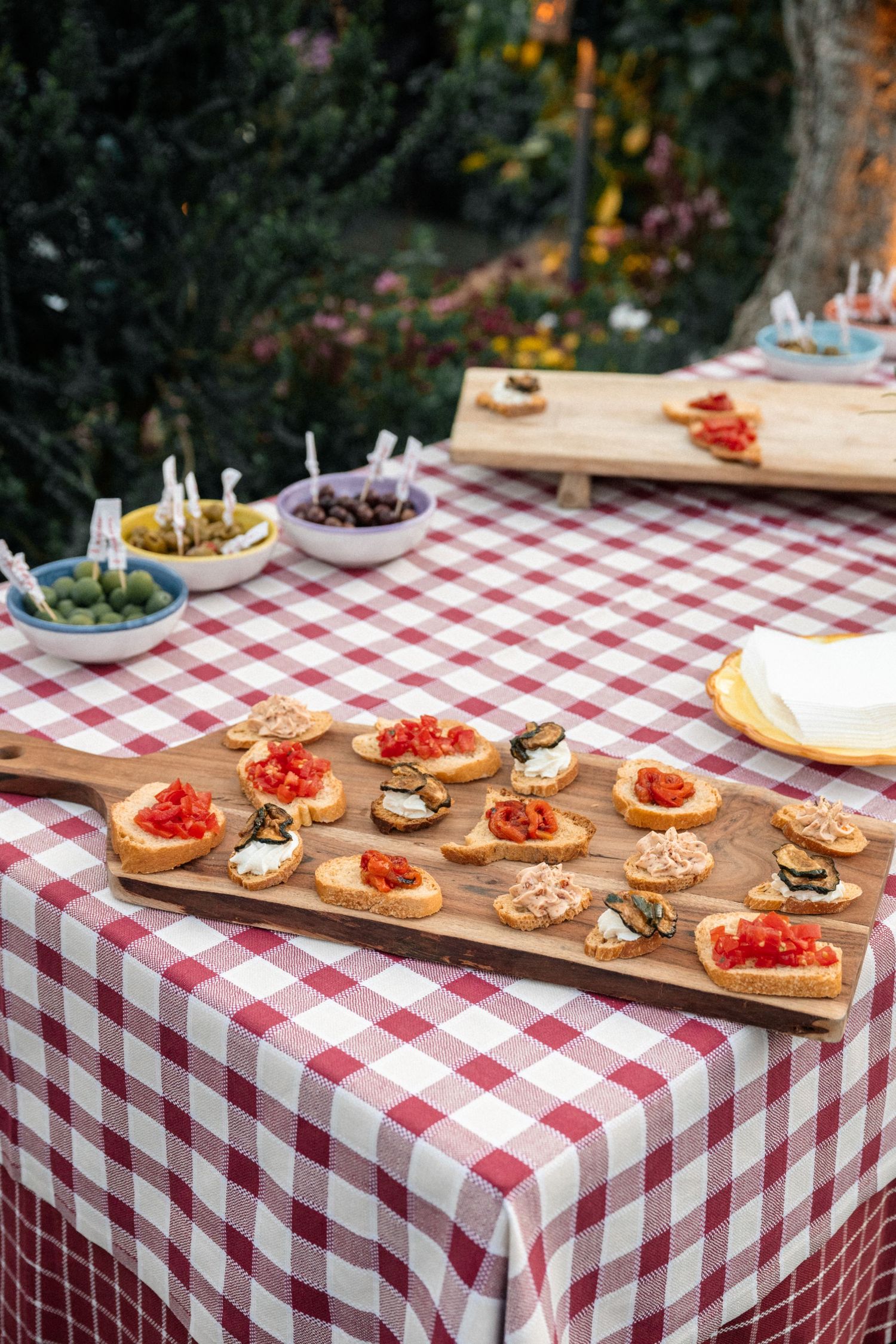 Wooden board of assorted bruschetta appetizers on red gingham tablecloth at an outdoor Italian wedding.