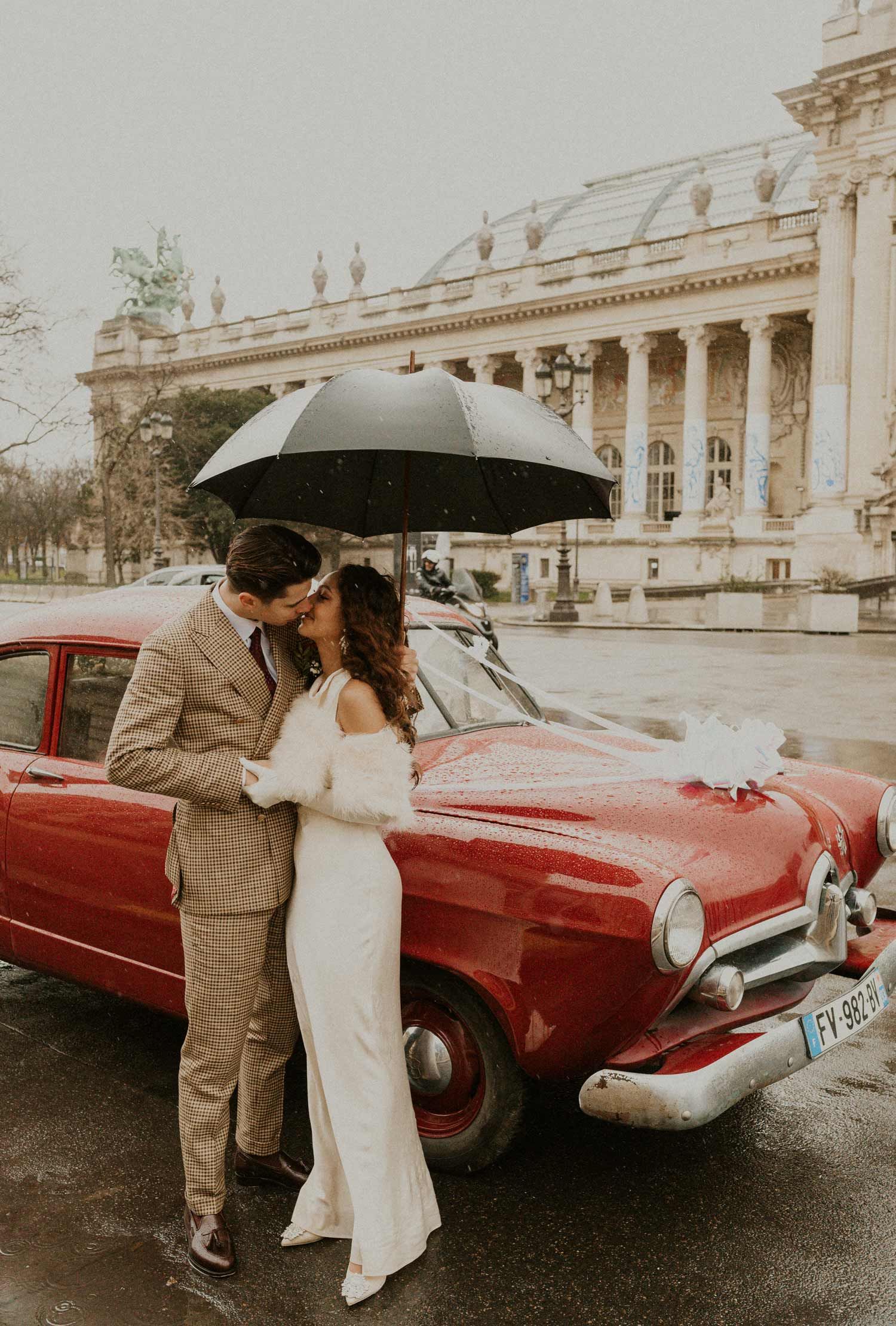 A couple poses with a classic red car in front of a historic building on their wedding day.