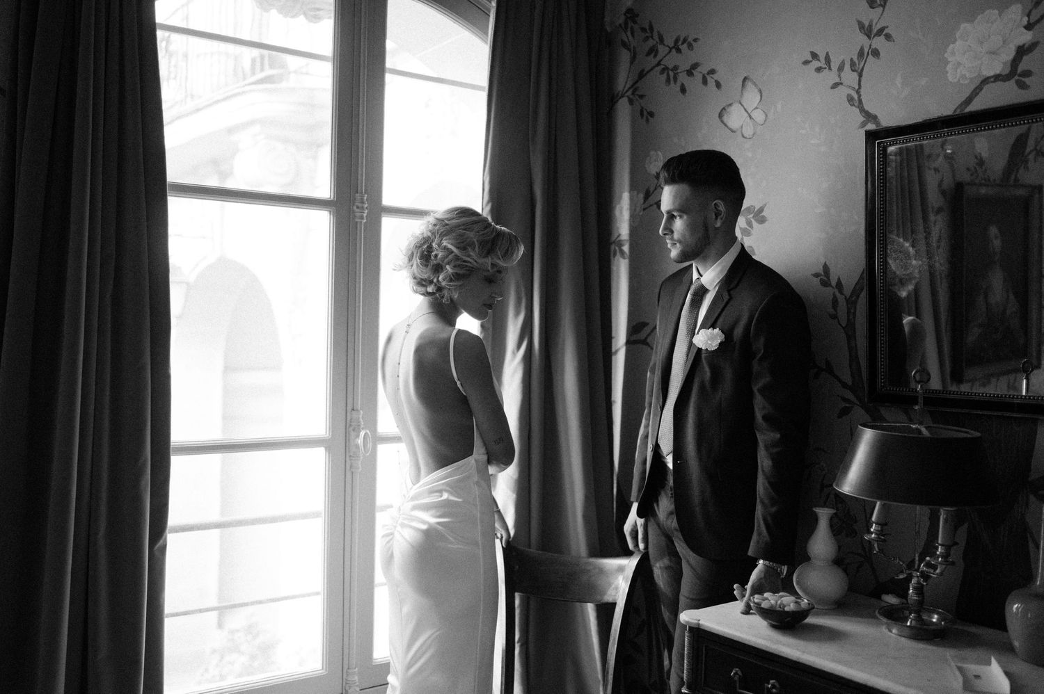 Black and white photo of couple by window in elegant room with ornate moldings.