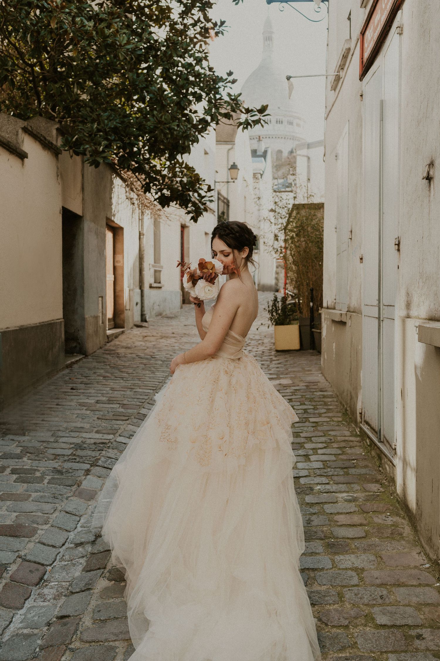 A bride wearing a romantic tulle gown stands in a quaint cobblestone alley during golden hour.