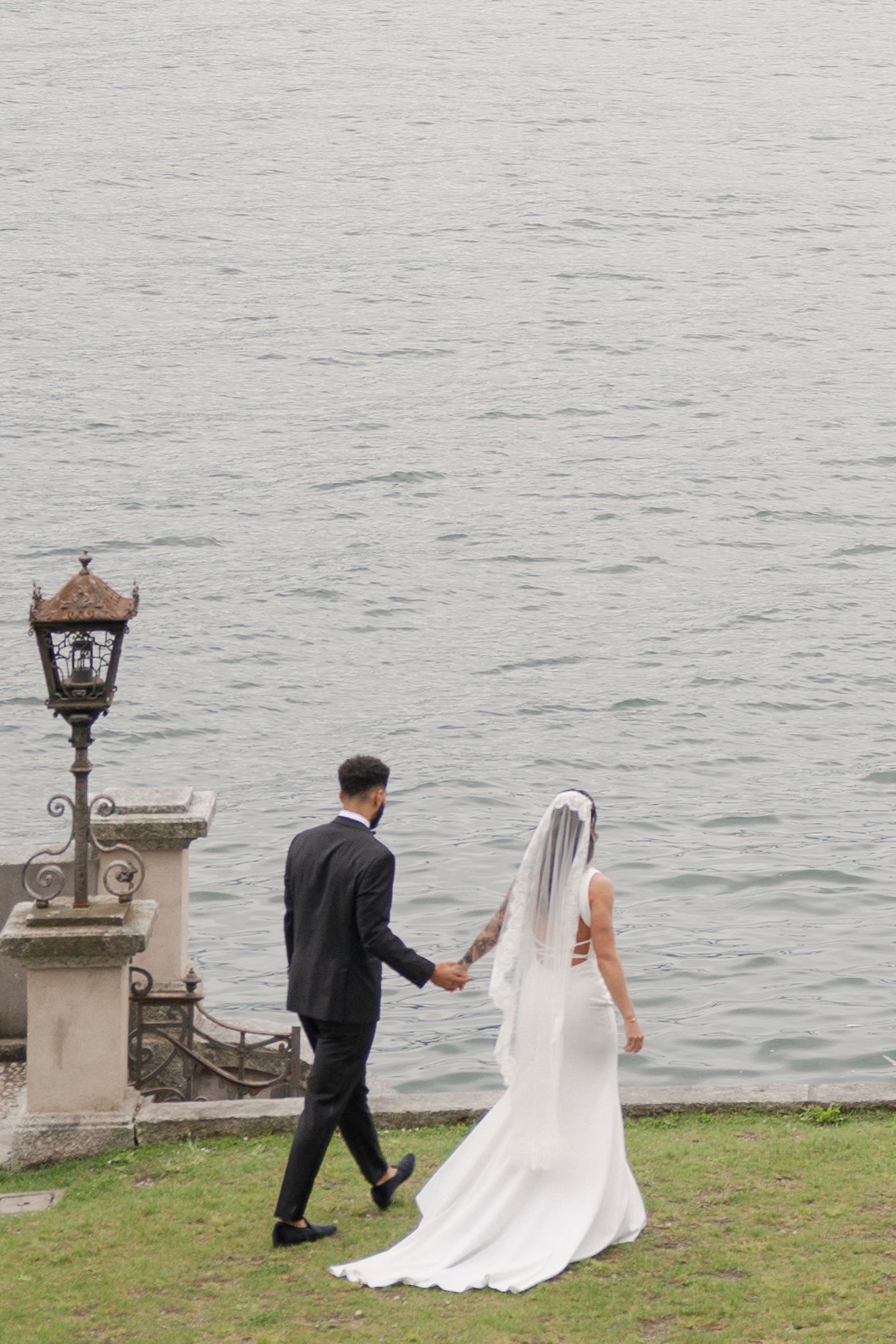 Bride and groom holding hands walking toward a serene lake, bride in white gown with veil, groom in black suit.