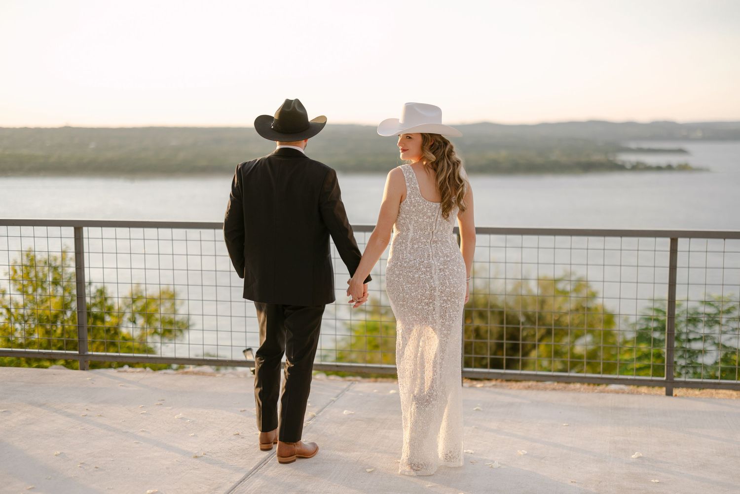 Couple in western attire holding hands overlooking a scenic lake from an outdoor terrace at golden hour.