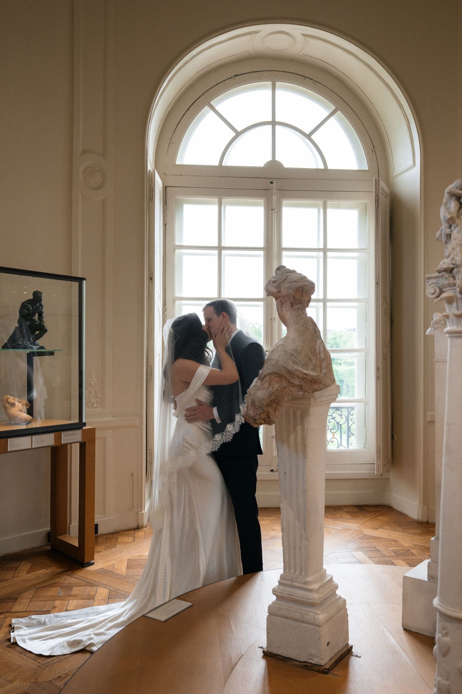 Bride and groom sharing a kiss by arched window with classical sculptures.