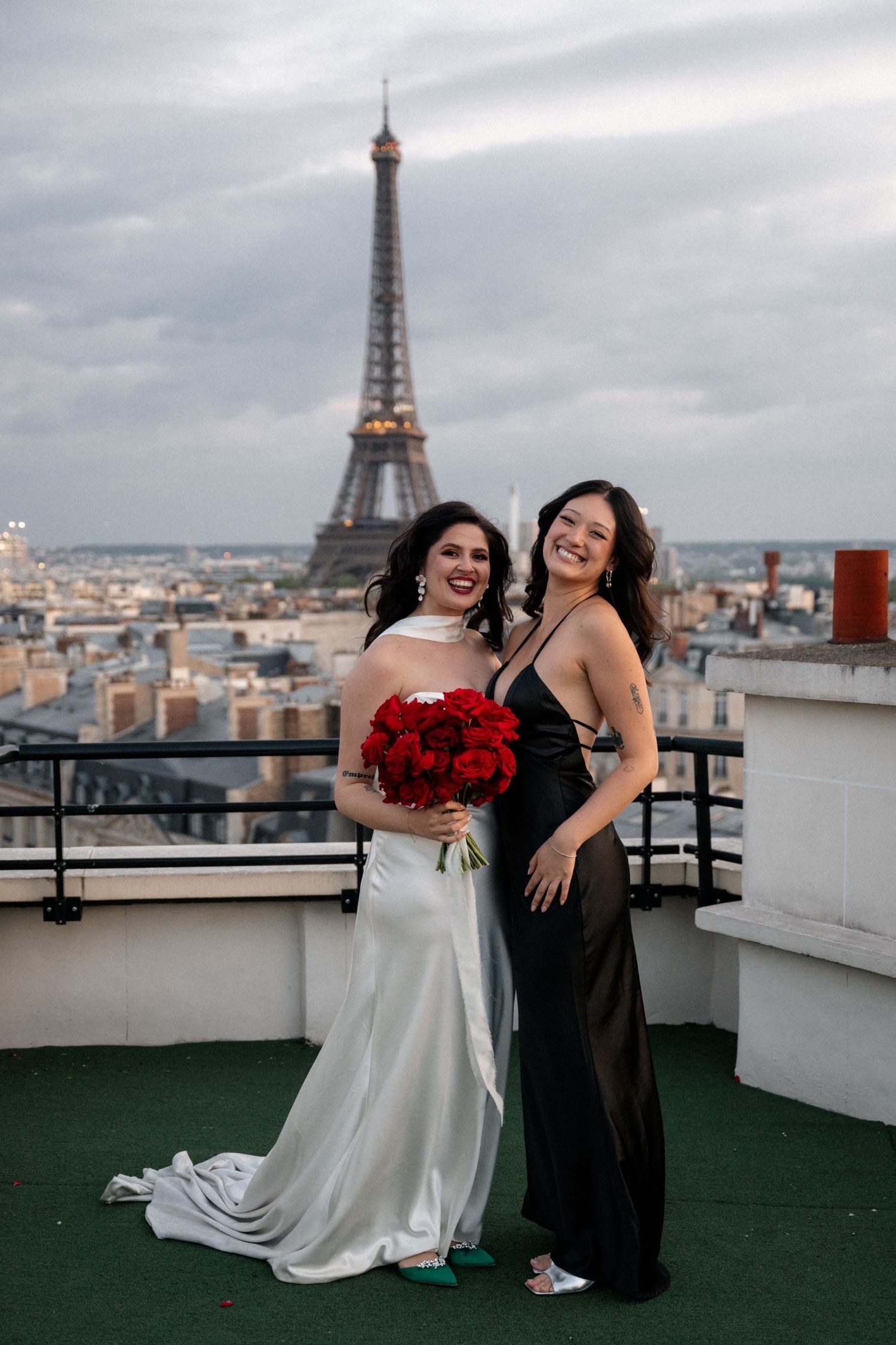 Bride in white satin gown with bridesmaid in black dress posing before the Eiffel Tower.