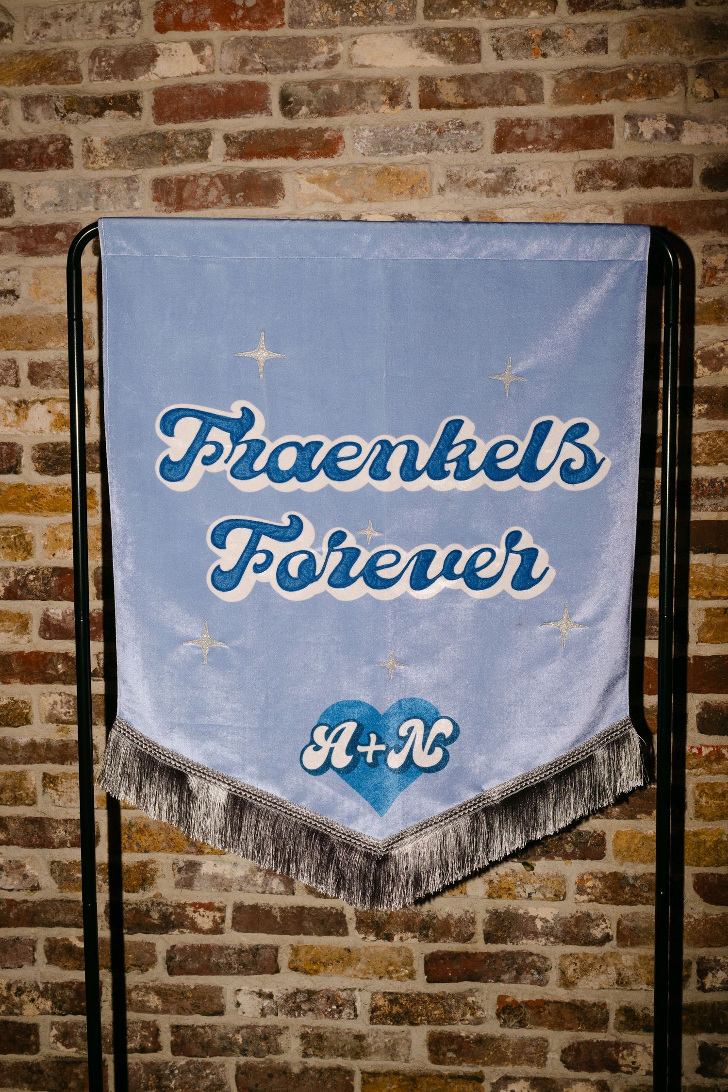Light blue velvet banner reading Fraenkels Forever with initials A+N in a blue heart, displayed on a metal stand against brick wall.