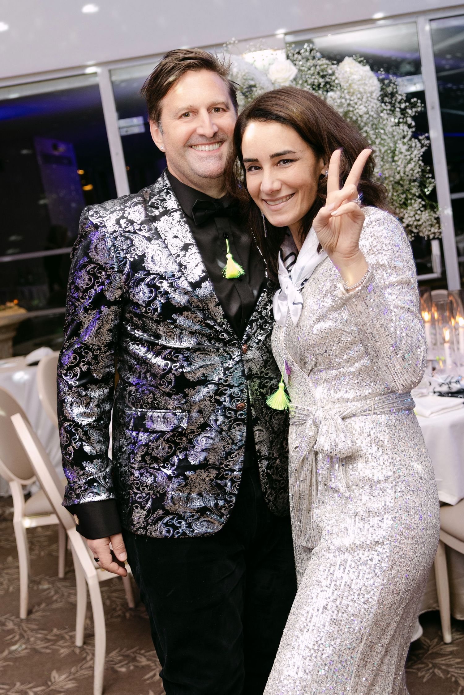 Couple posing at formal event, man in floral blazer, woman in silver sequin dress flashing peace sign.