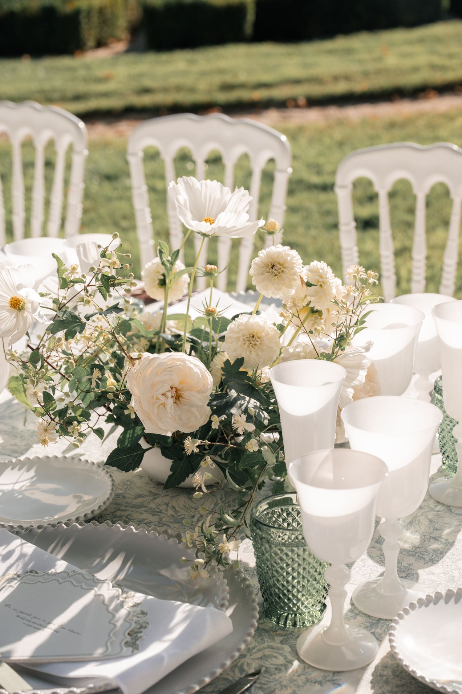 White floral centerpiece with milk glass goblets on elegant outdoor wedding table setting.