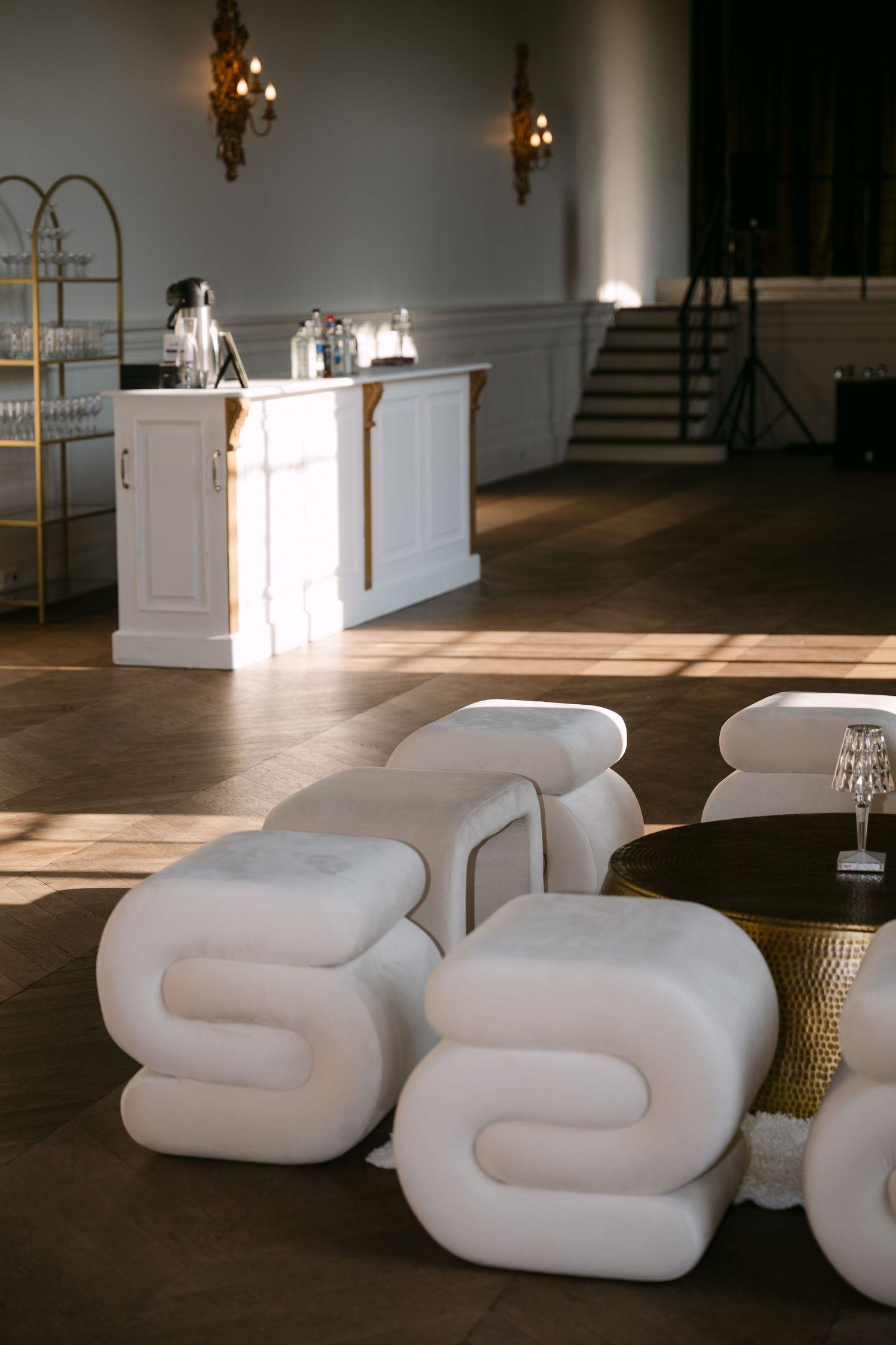 Chic event space with white sculptural lounge seating, gold accents, and a white bar in a sunlit ballroom.