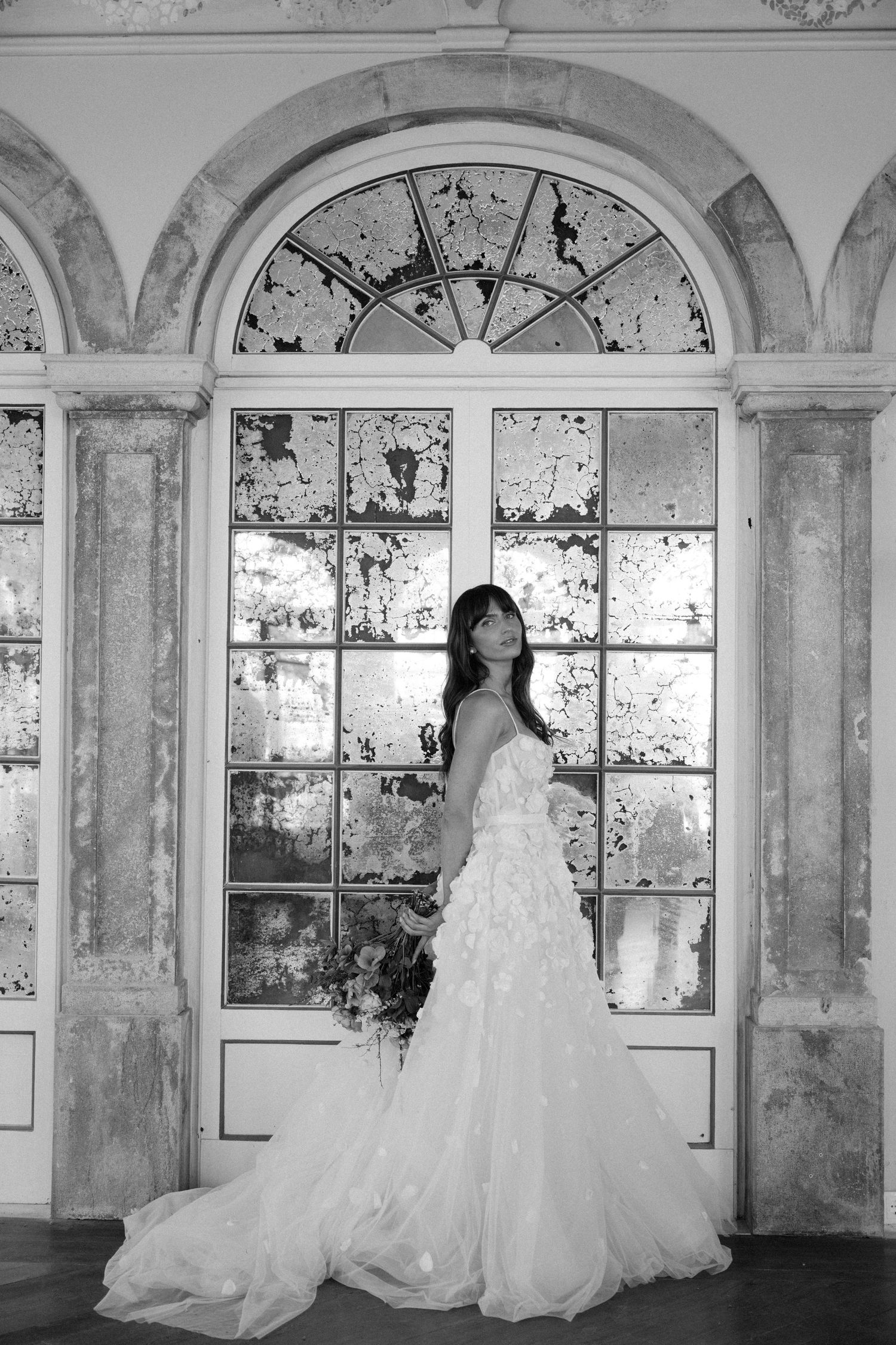 Elegant bride in a flowing white ruffled gown standing before ornate arched windows in black and white photo.