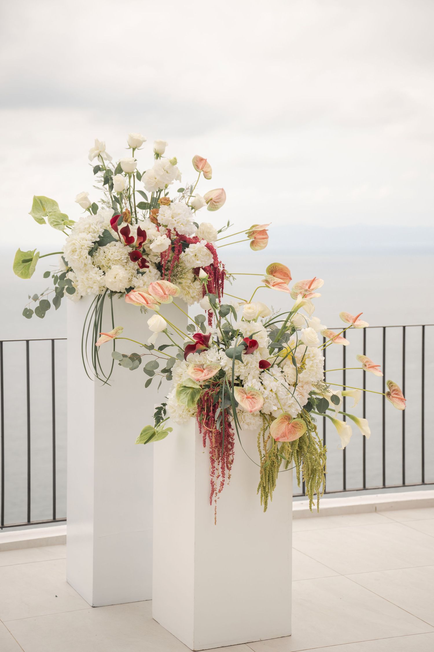 Two white pedestals topped with lush floral arrangements of white, pink, and red blooms on an outdoor terrace.