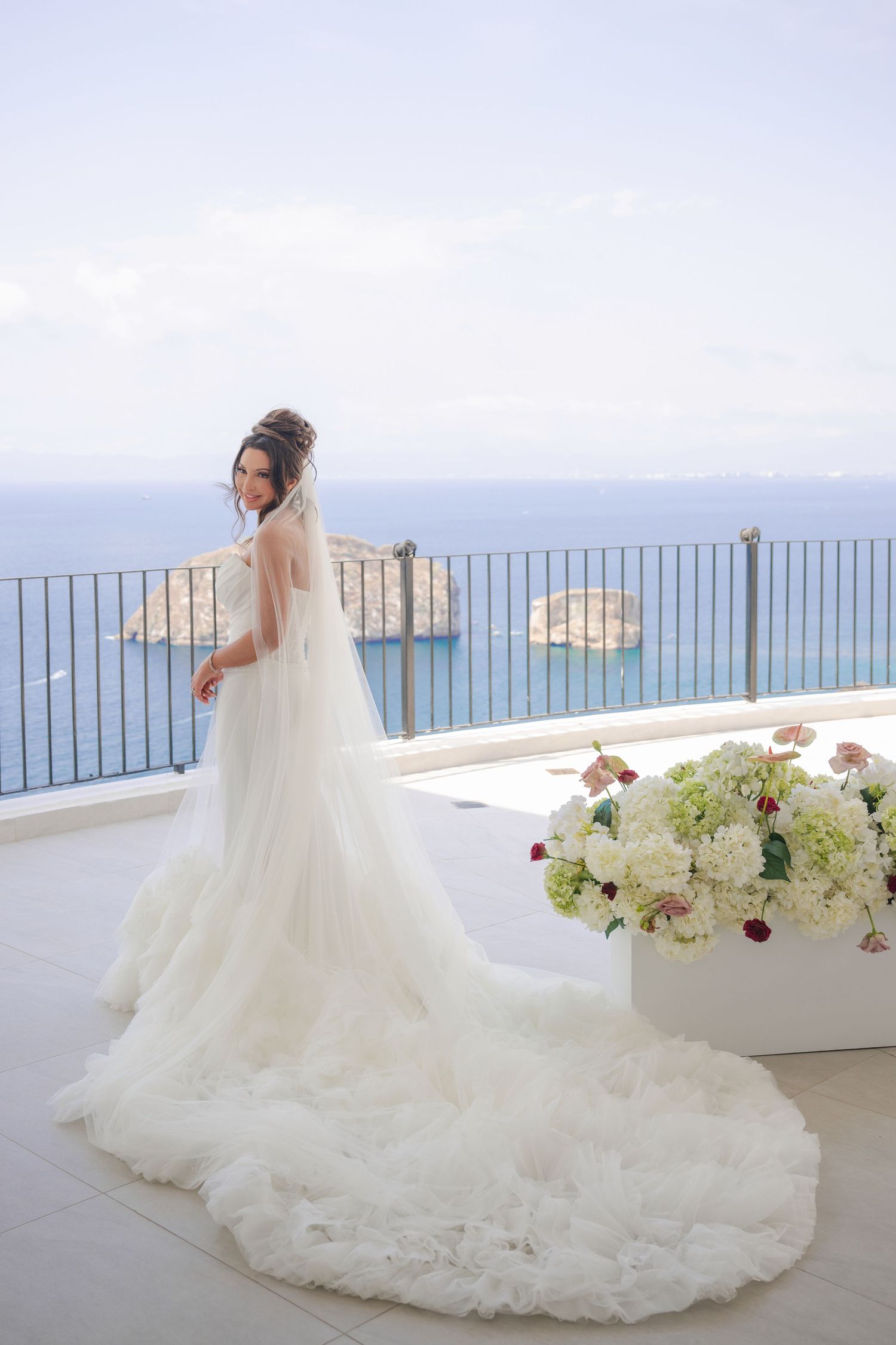 Bride in a stunning white gown with long veil stands on a terrace with breathtaking ocean views and white floral decor.