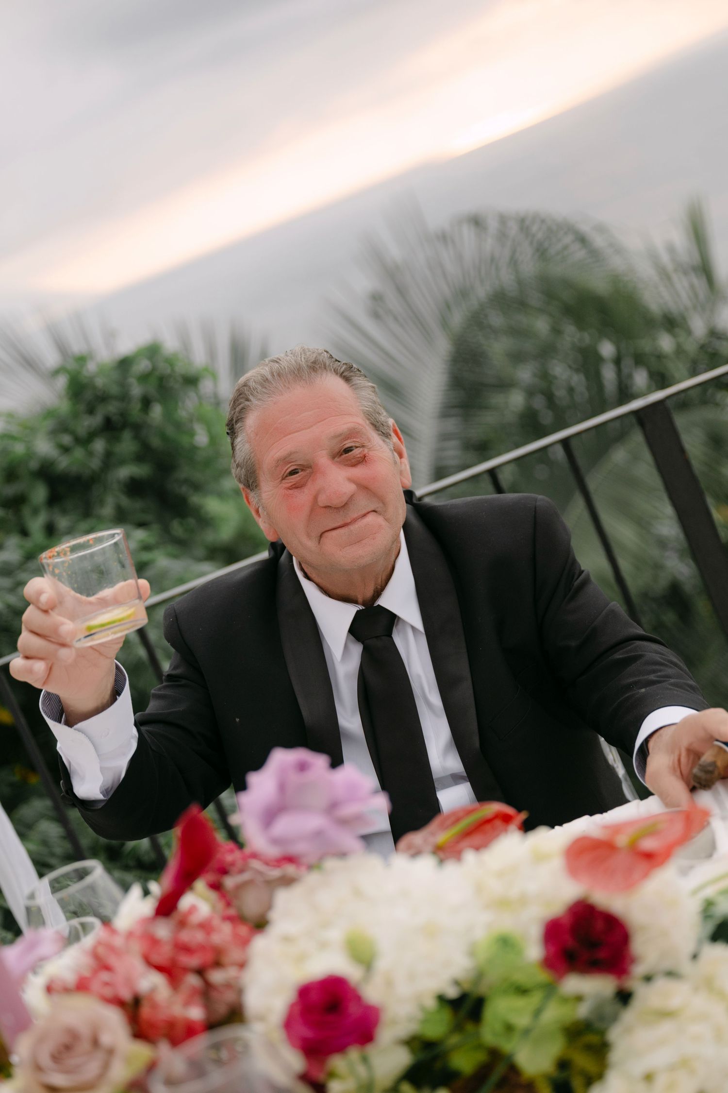Older man in black tuxedo raising a glass of champagne at an elegant outdoor wedding reception.