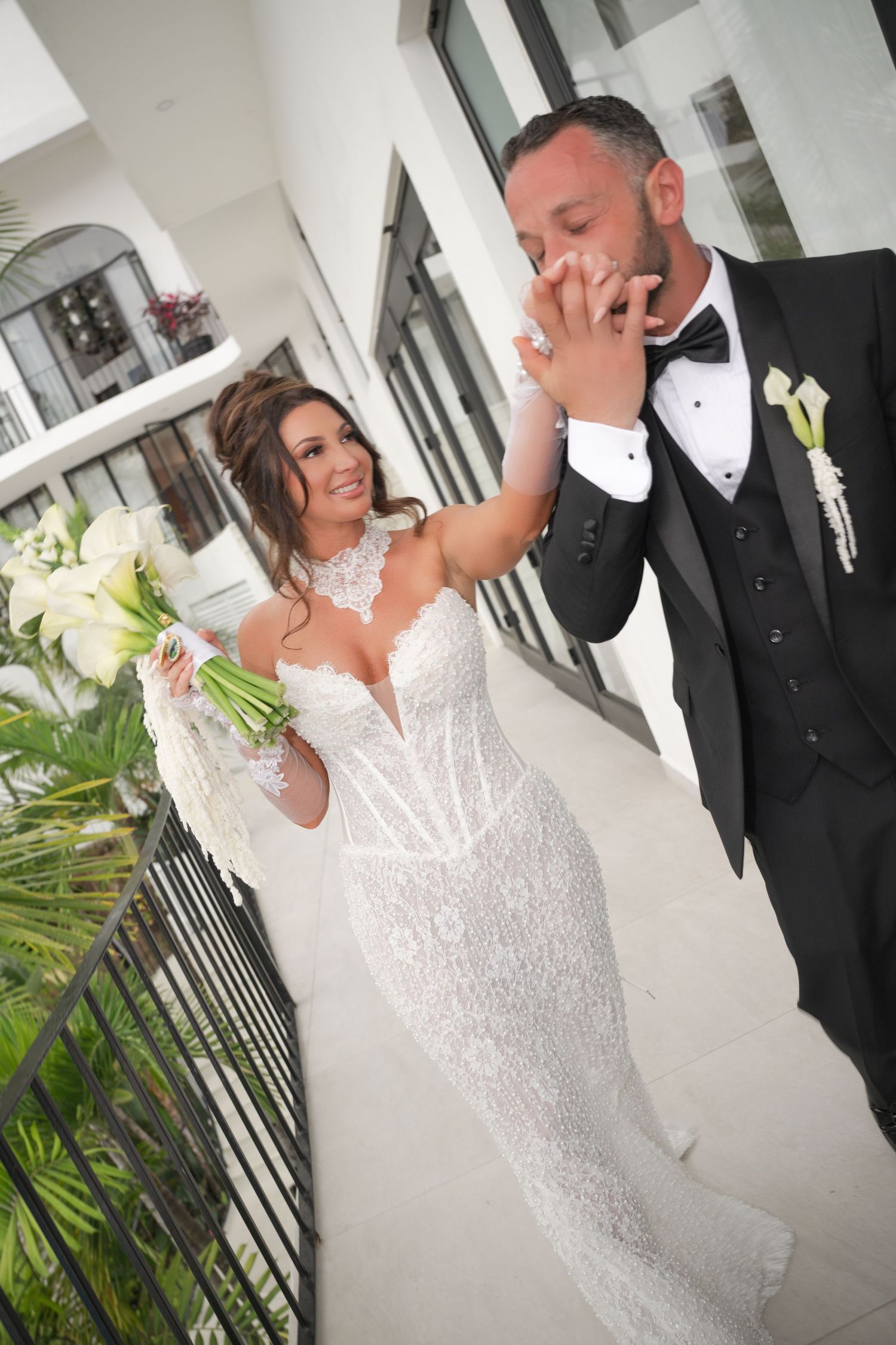 A groom in a tuxedo kisses his bride's hand as she smiles holding a white bouquet.