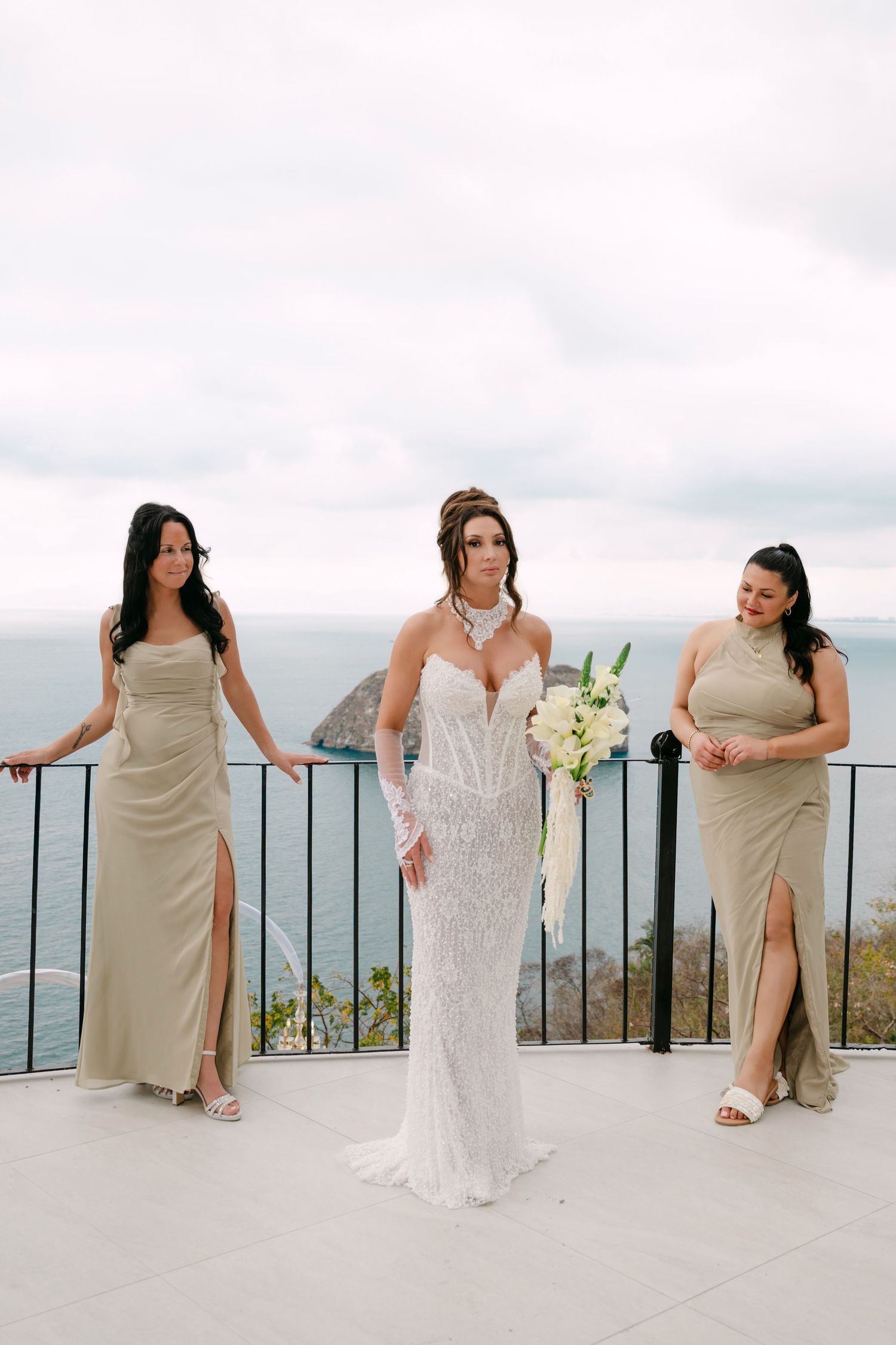 Bride in white beaded gown holds bouquet flanked by bridesmaids in gold dresses on scenic ocean overlook terrace.