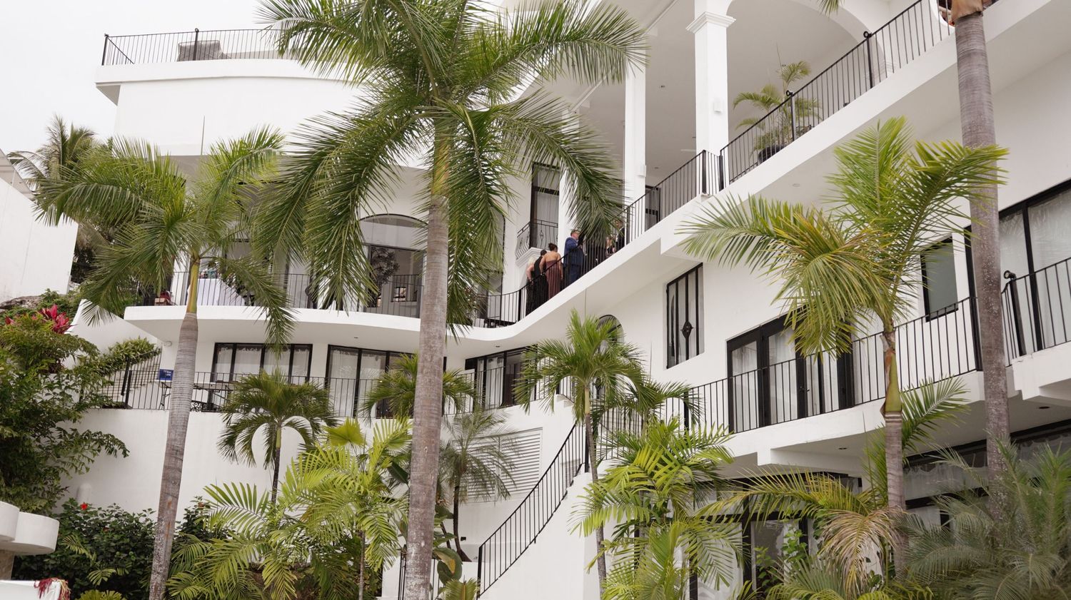 White multi-story hotel with palm trees, balconies, and guests enjoying tropical resort surroundings.