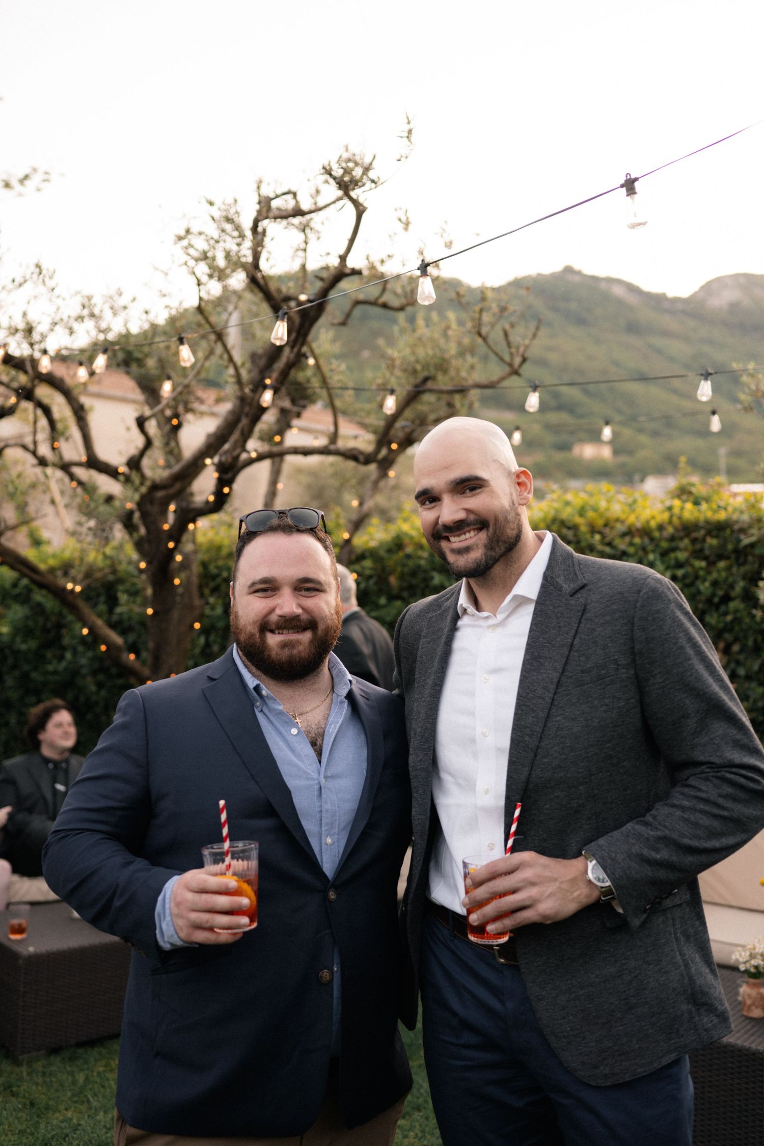 Two smiling men in blazers holding Aperol Spritz cocktails at an outdoor wedding reception in Italy.