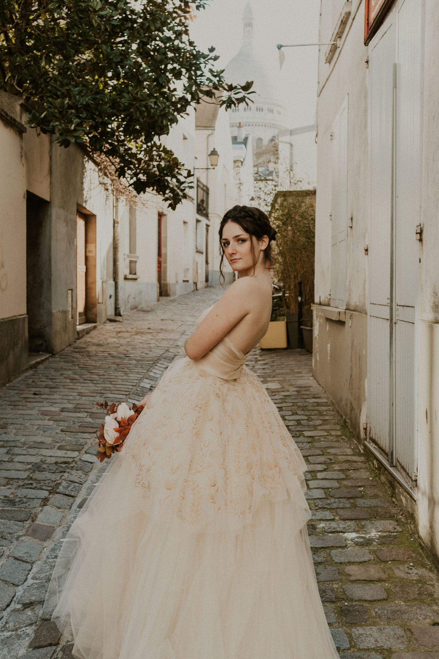 A bride in a champagne-colored ballgown poses on a cobblestone street with historic white buildings in the background.