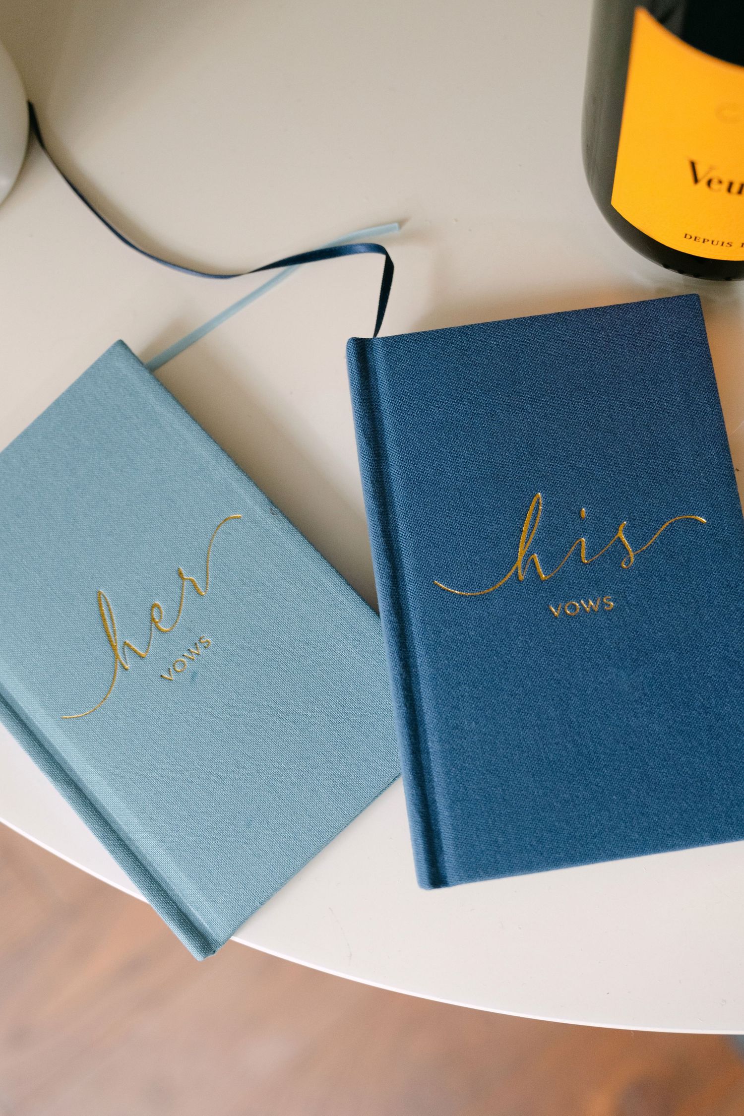 His and her wedding vow books in light blue and navy with gold lettering on a white surface.
