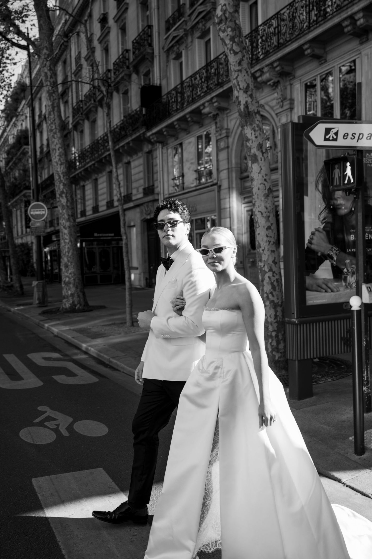 A couple in white wedding attire walks together through a sunlit European city street with classic architecture.