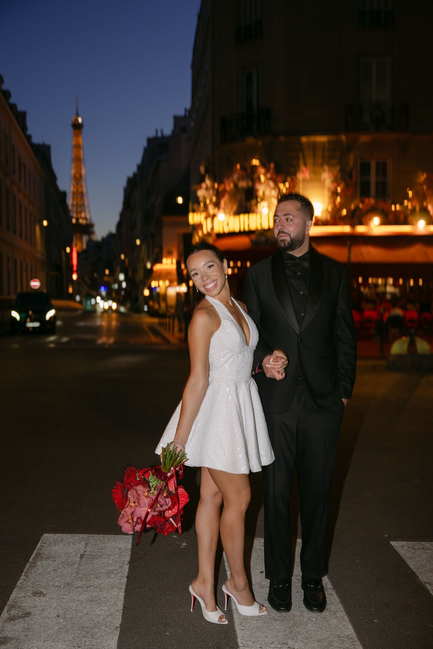 Couple in wedding attire posing on Paris street at night with Eiffel Tower glowing in background.
