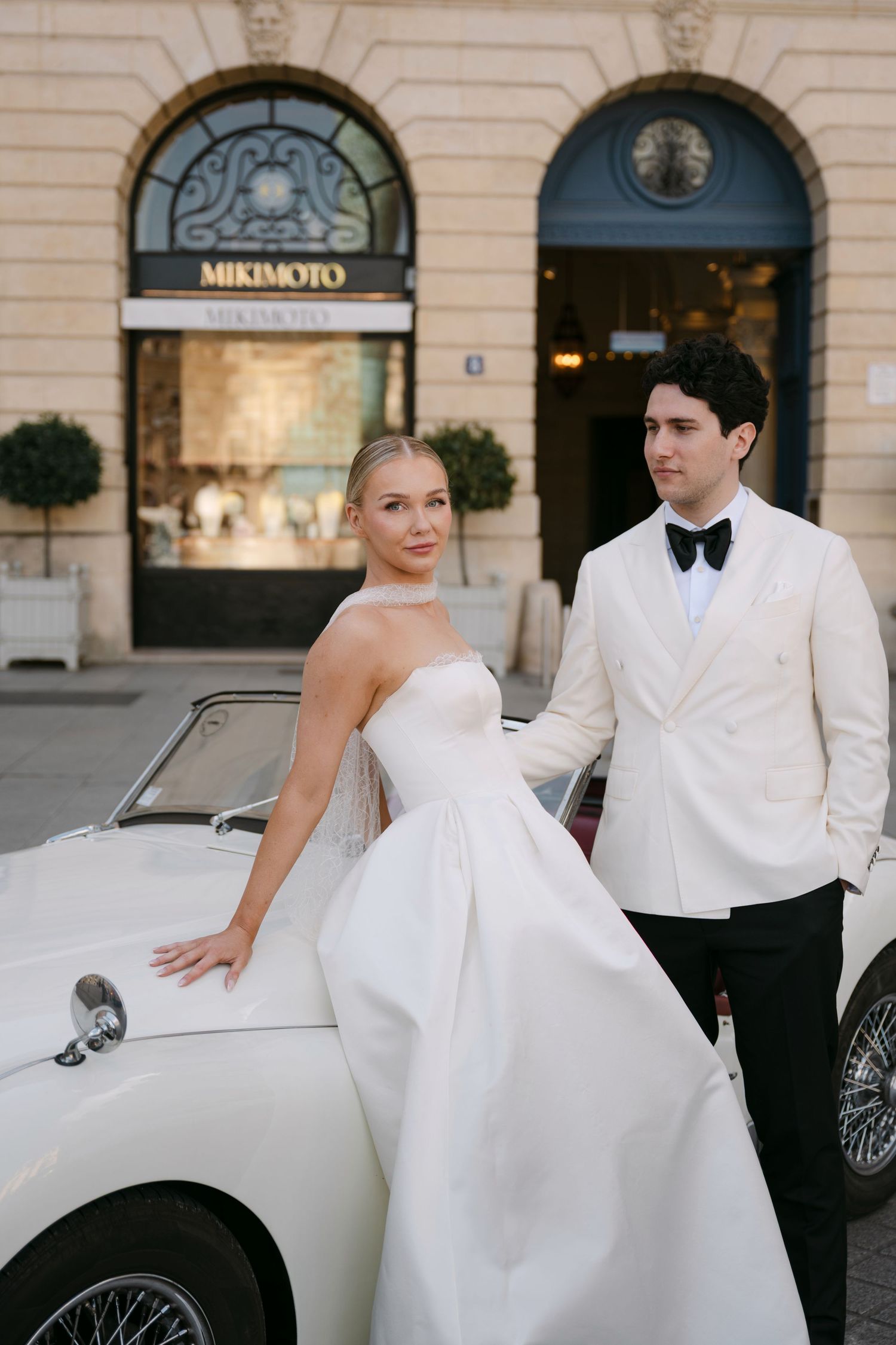 Bride in strapless white gown and groom in ivory tuxedo pose by vintage white convertible outside Mikimoto store.