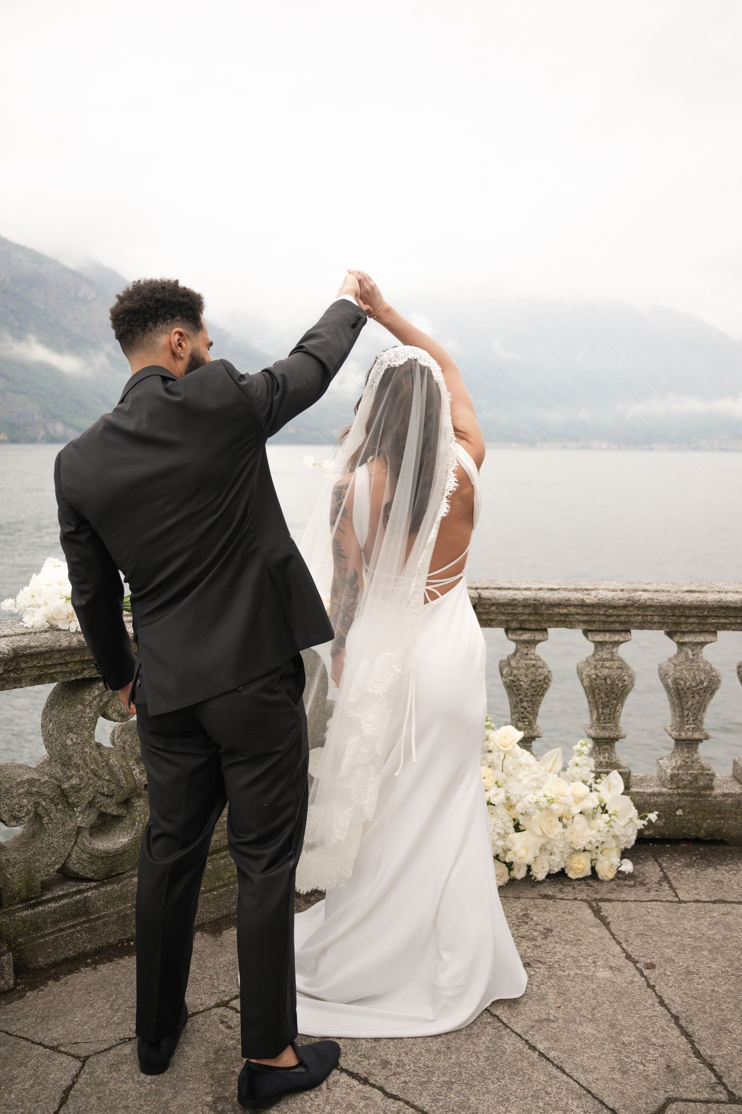Groom spinning bride in white gown and veil on a stone balcony overlooking a misty lake.