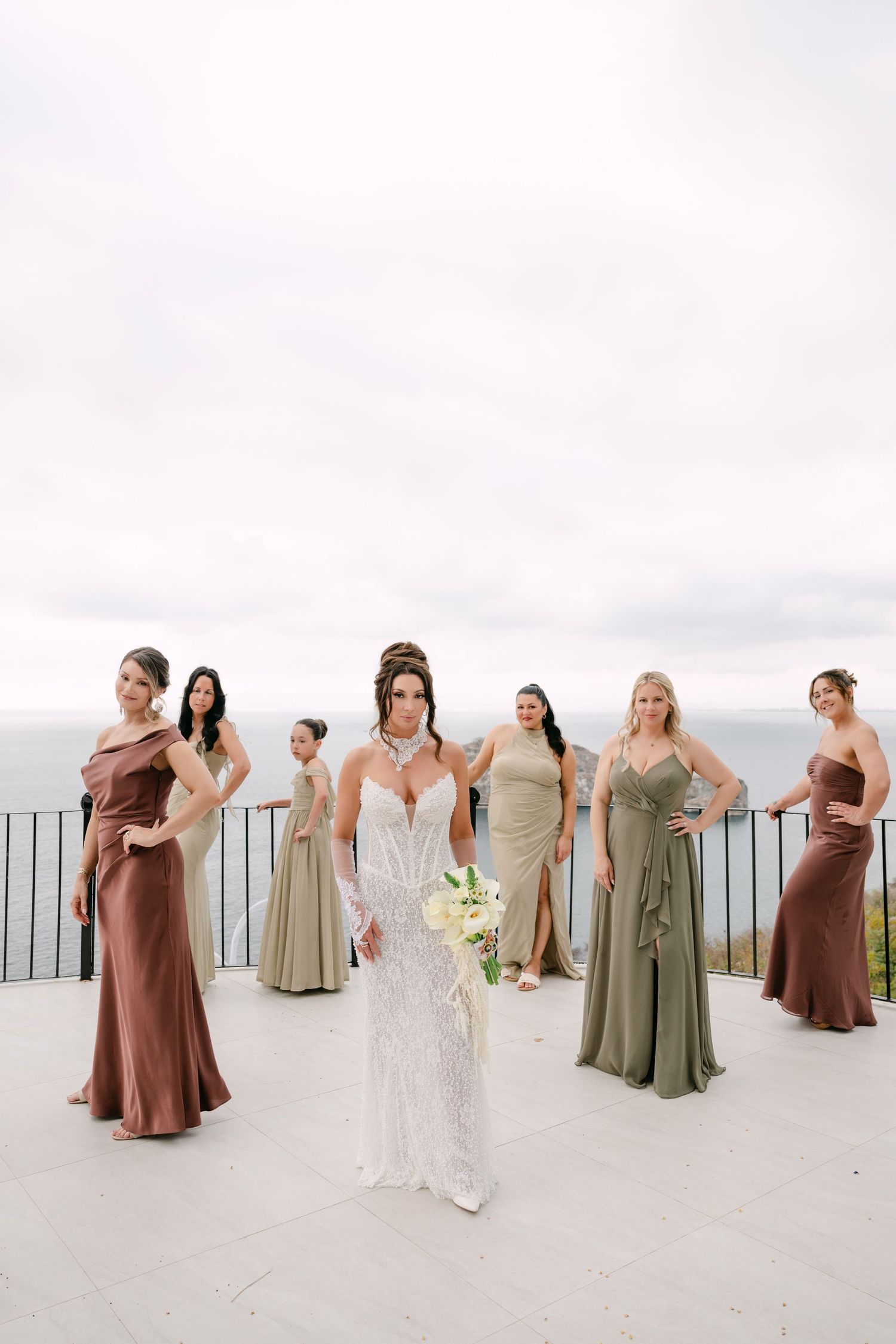 Bride in white lace gown holds bouquet with bridesmaids in earth-tone satin dresses on ocean-view terrace.