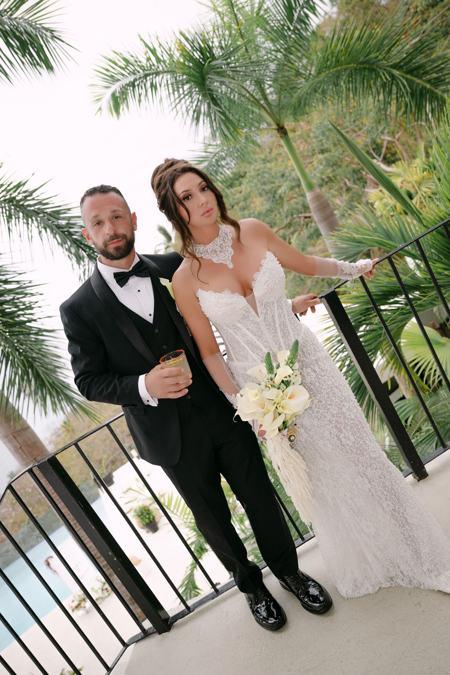 Bride and groom pose on balcony surrounded by palm trees, she in lace gown, he in black tuxedo holding champagne.