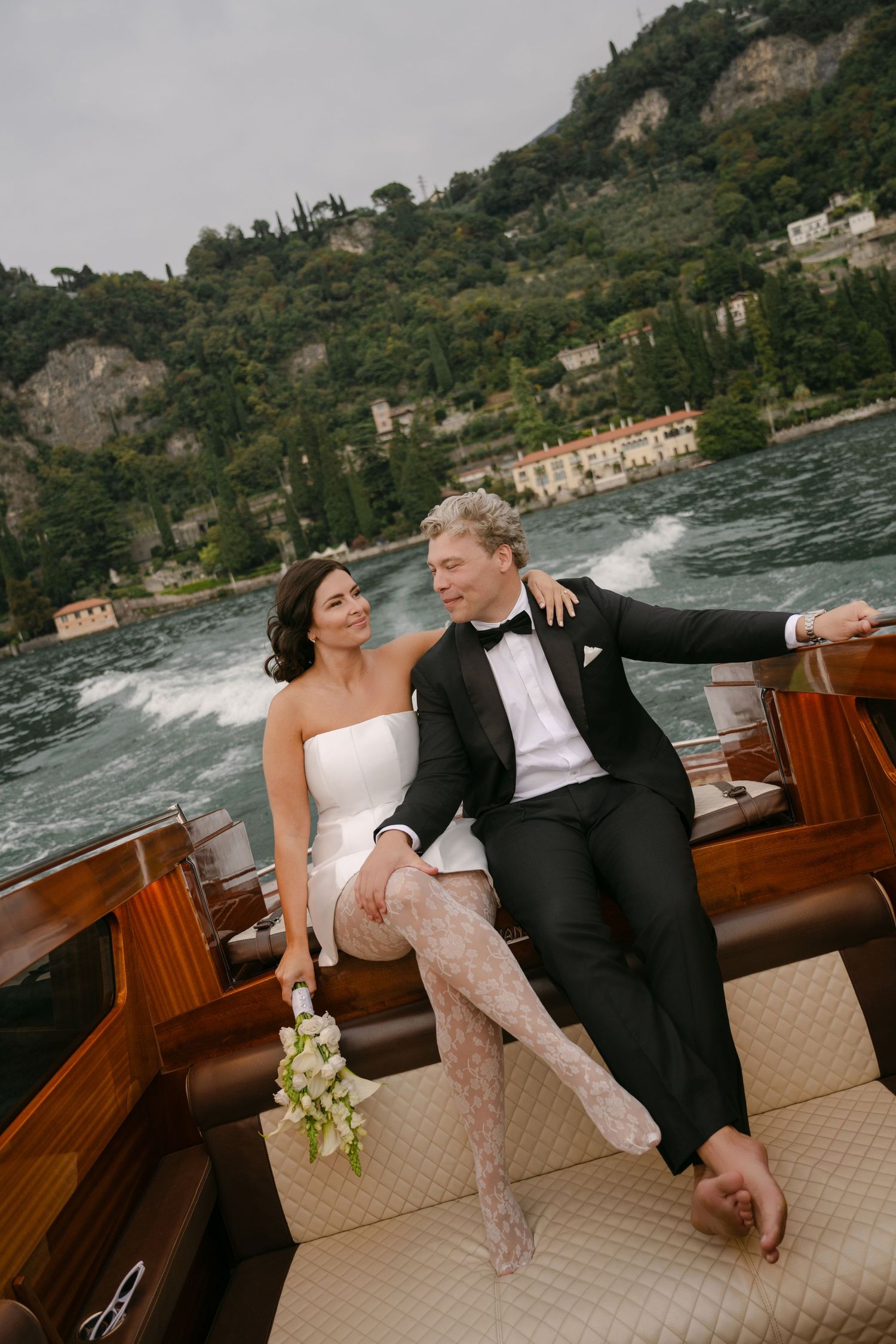 Bride in strapless white dress and groom in black tuxedo sit close on a vintage wooden boat cruising a scenic lakeside with hillside villas.